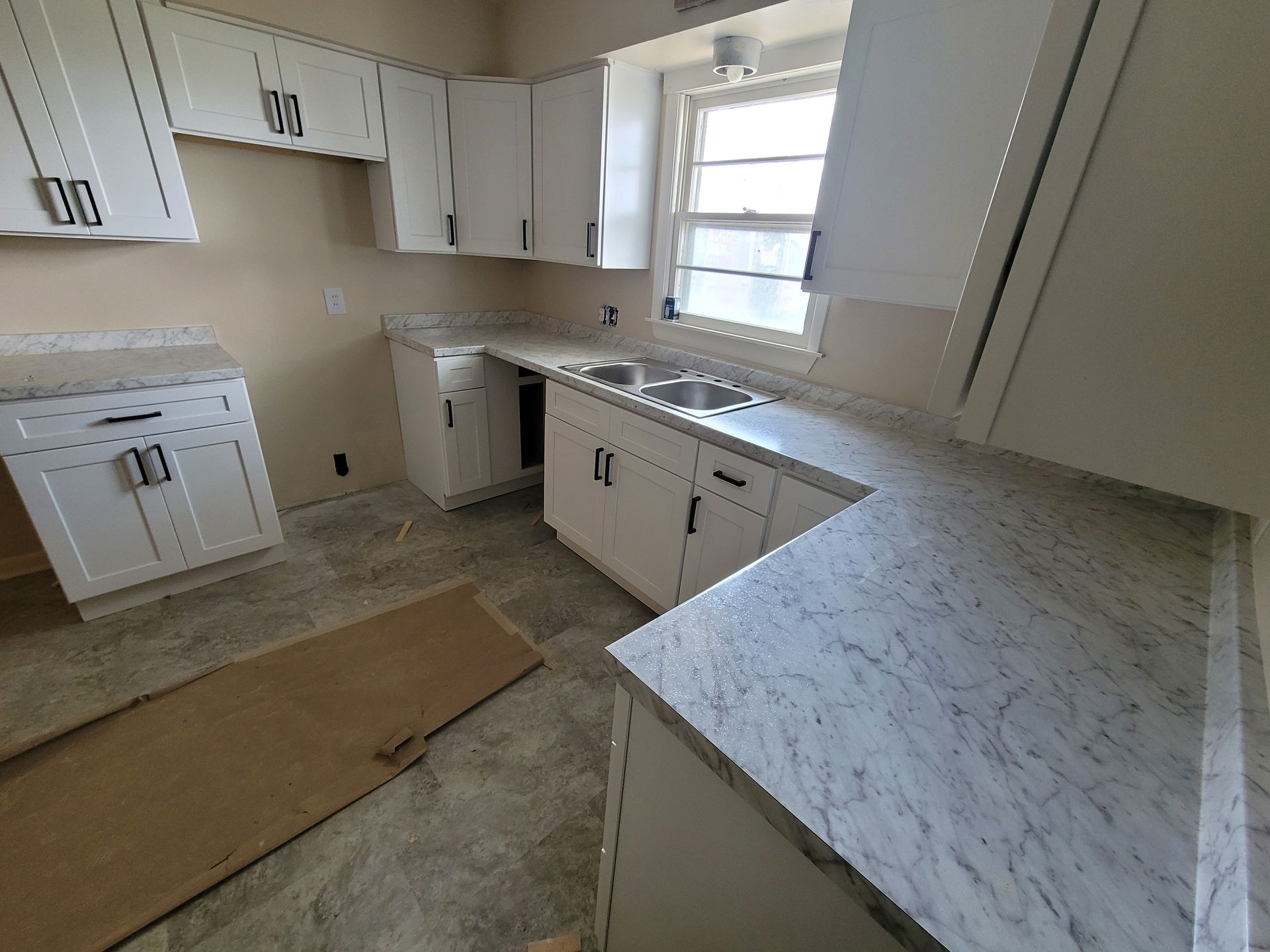 White kitchen cabinets and countertops in a room with a window; unfinished flooring.
