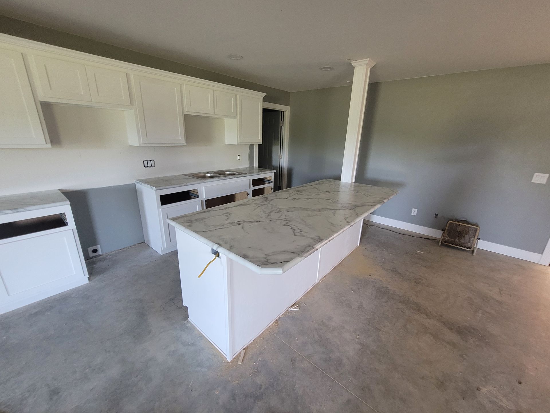 White kitchen cabinets and island with grey countertops in a room with grey walls and a concrete floor.