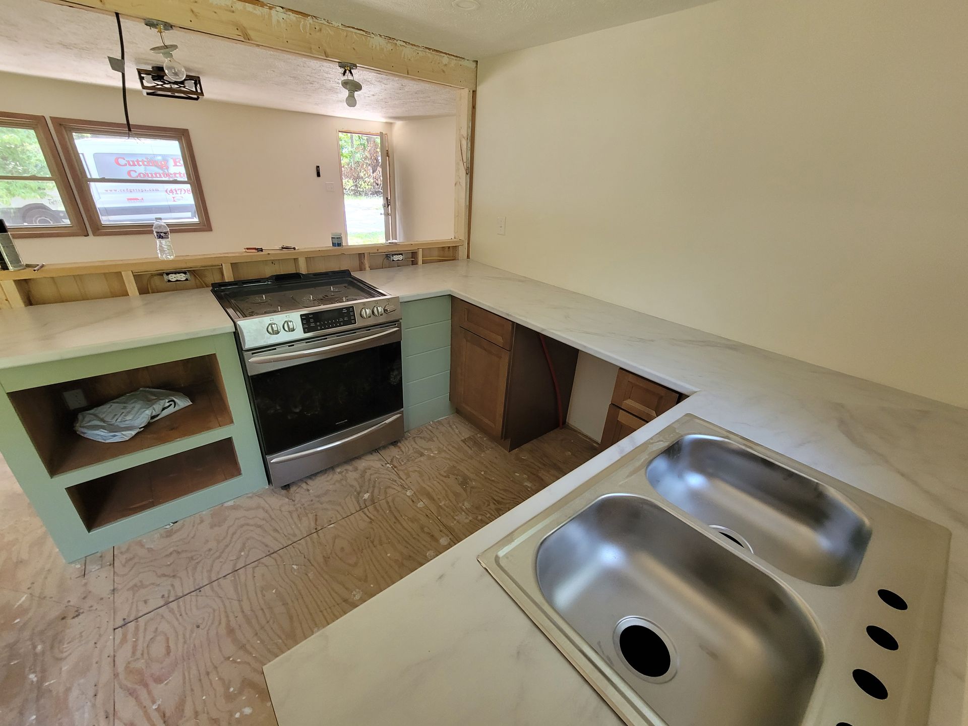 Kitchen renovation in progress: a gas range, sink, and countertops installed. Green and brown cabinets. Bright room.