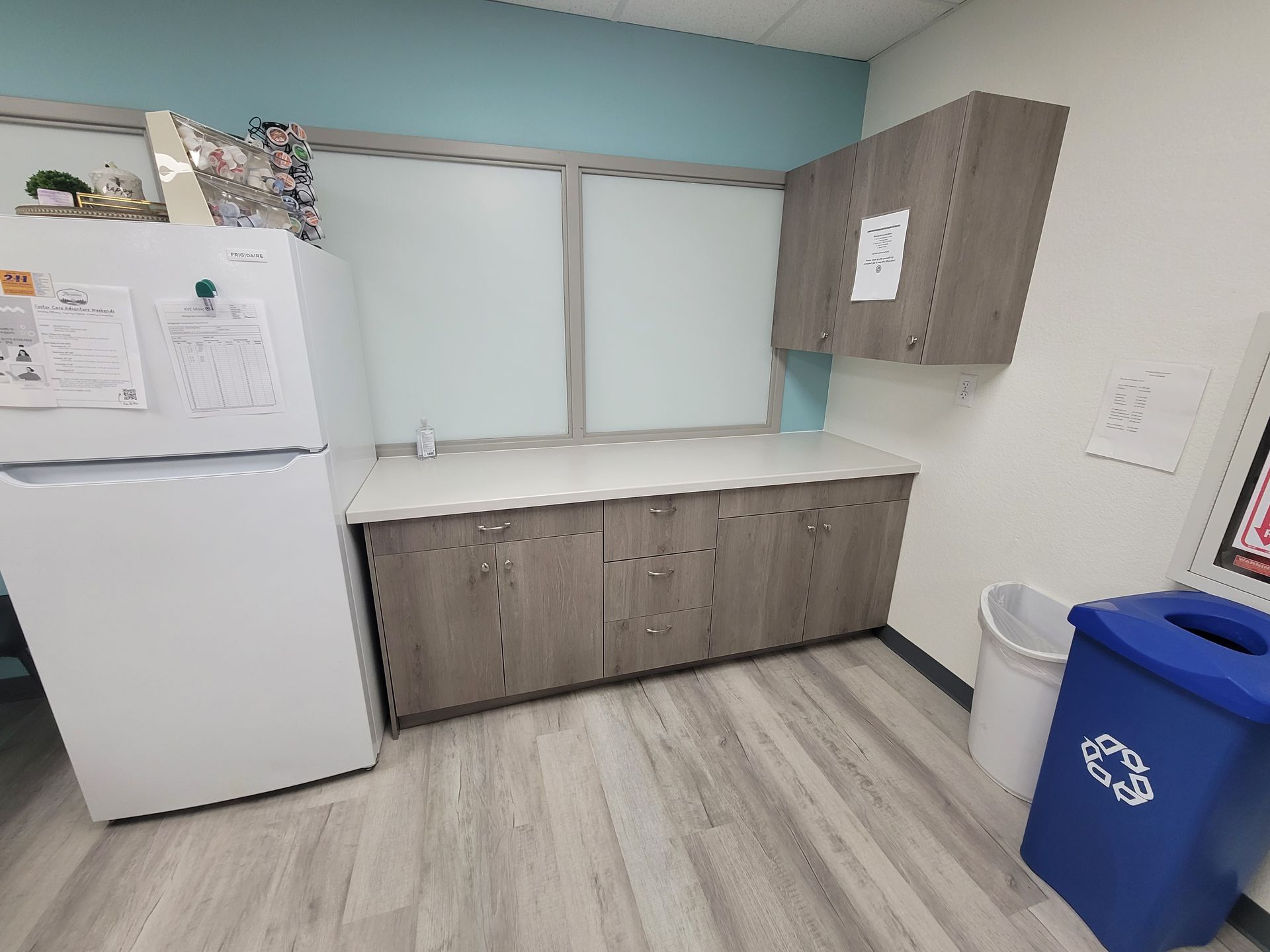 Kitchenette with cabinets, a fridge, and a recycling bin, set in a room with light blue walls and wooden floors.