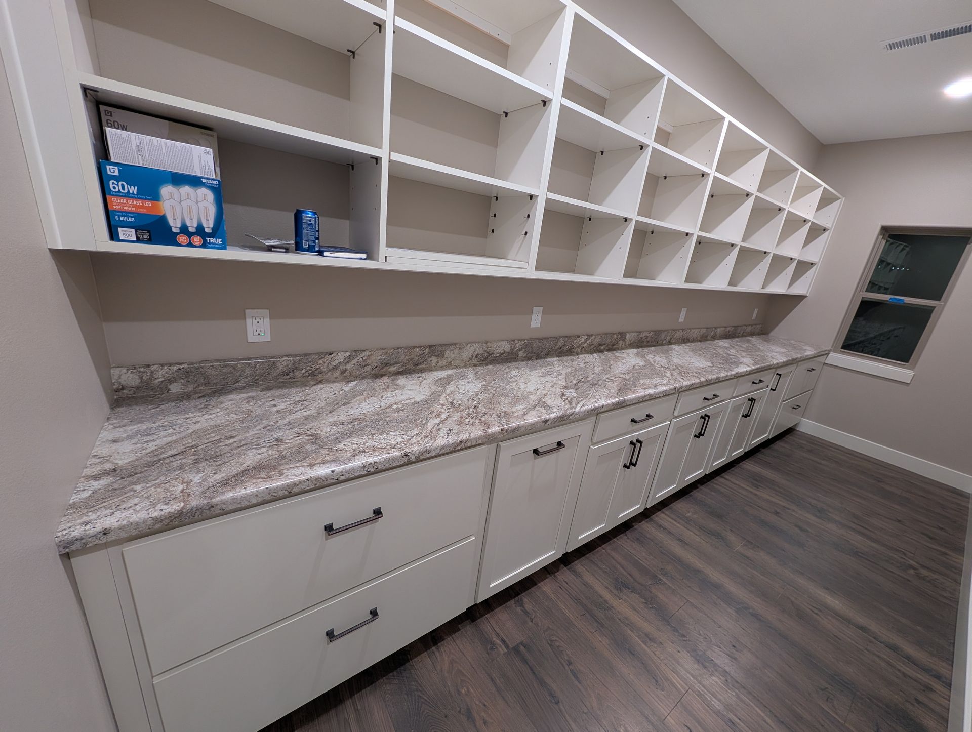 Pantry with white shelves, cabinets, and a granite countertop. Includes a window and wooden floor.