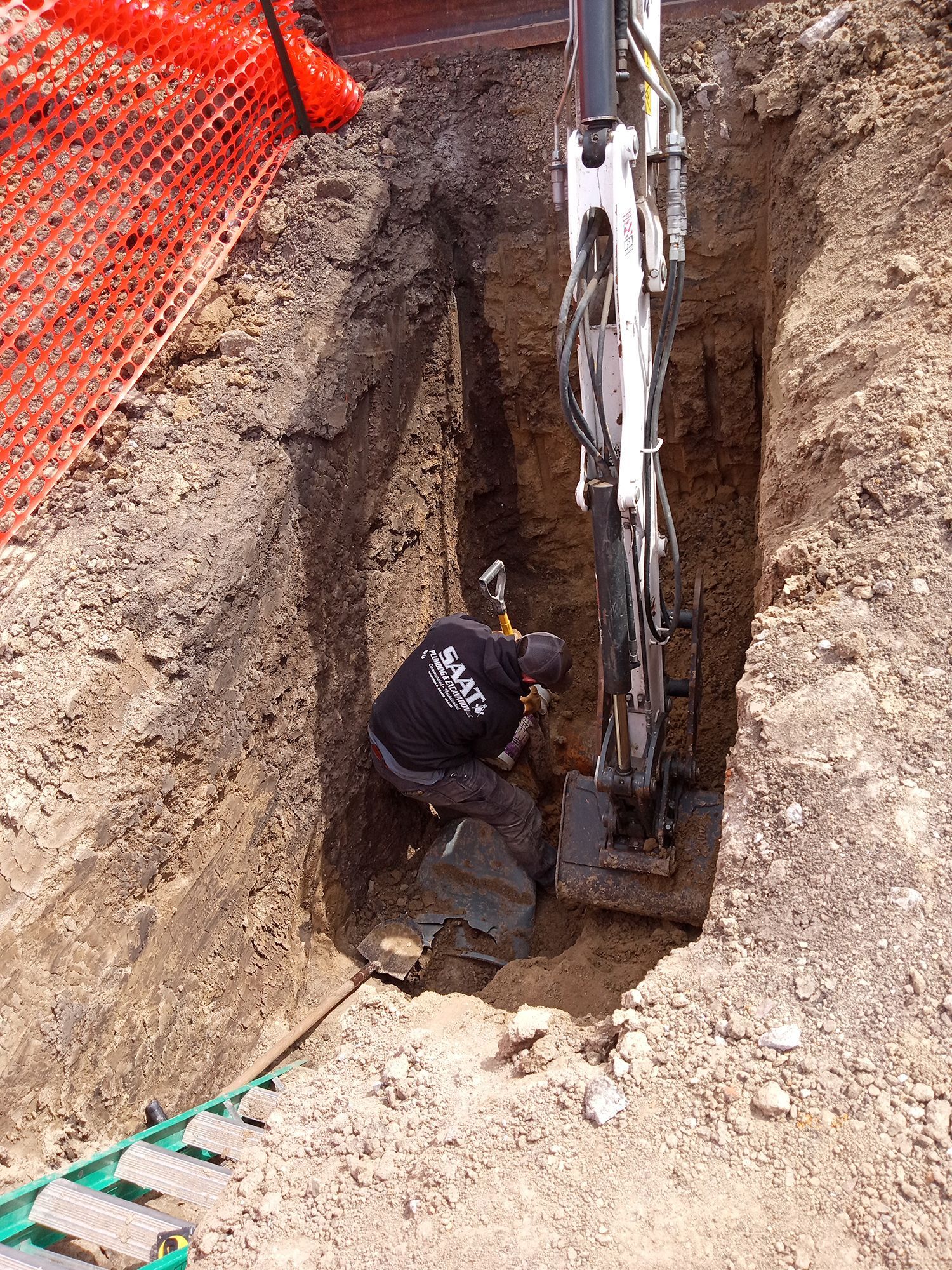 Worker in a trench, operating machinery. Dirt walls, safety fencing, and an excavator.