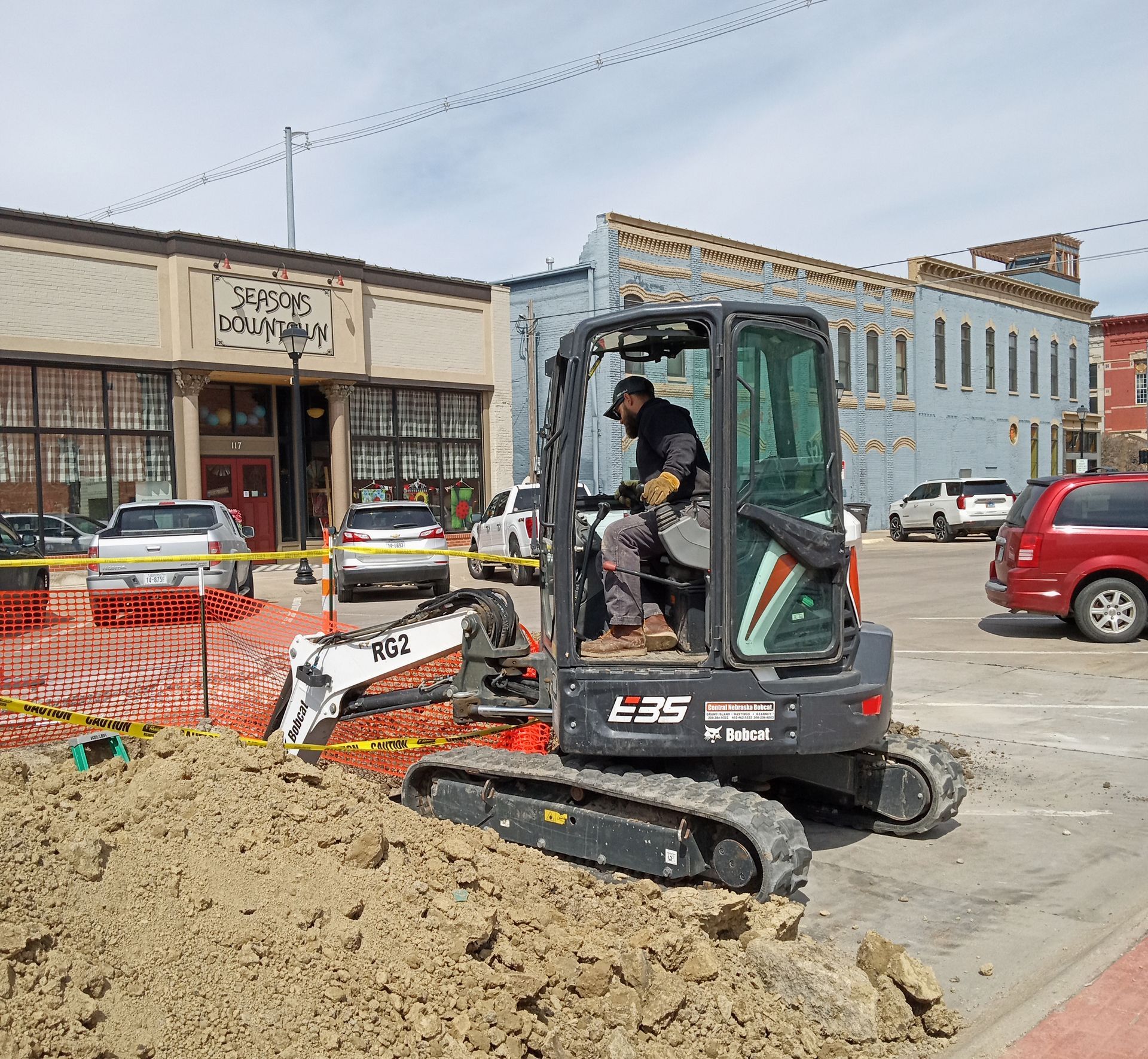 A person operating a Bobcat excavator on a city street; construction in progress near buildings and cars.