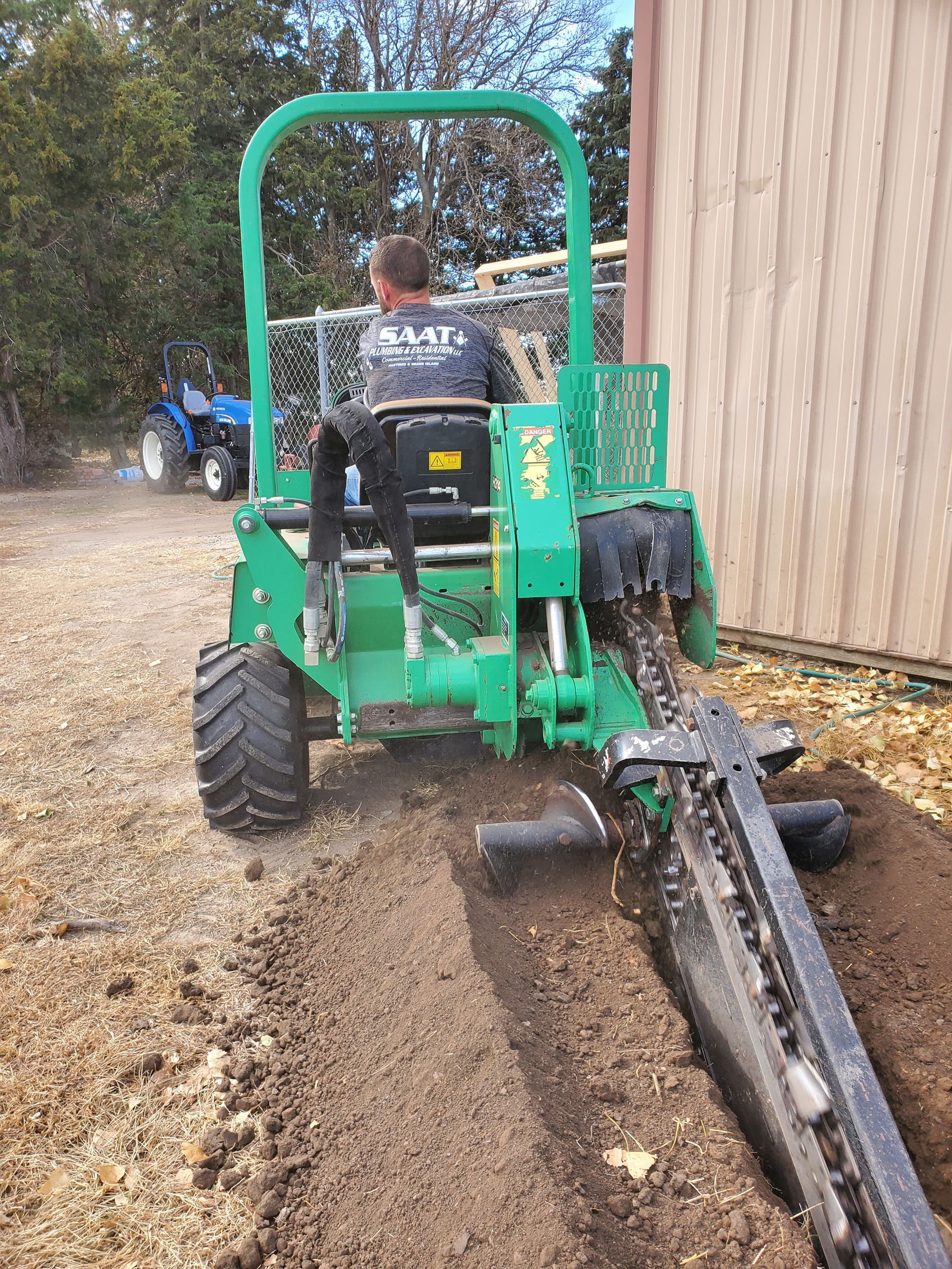 Man operating a green trencher, digging a trench next to a building.