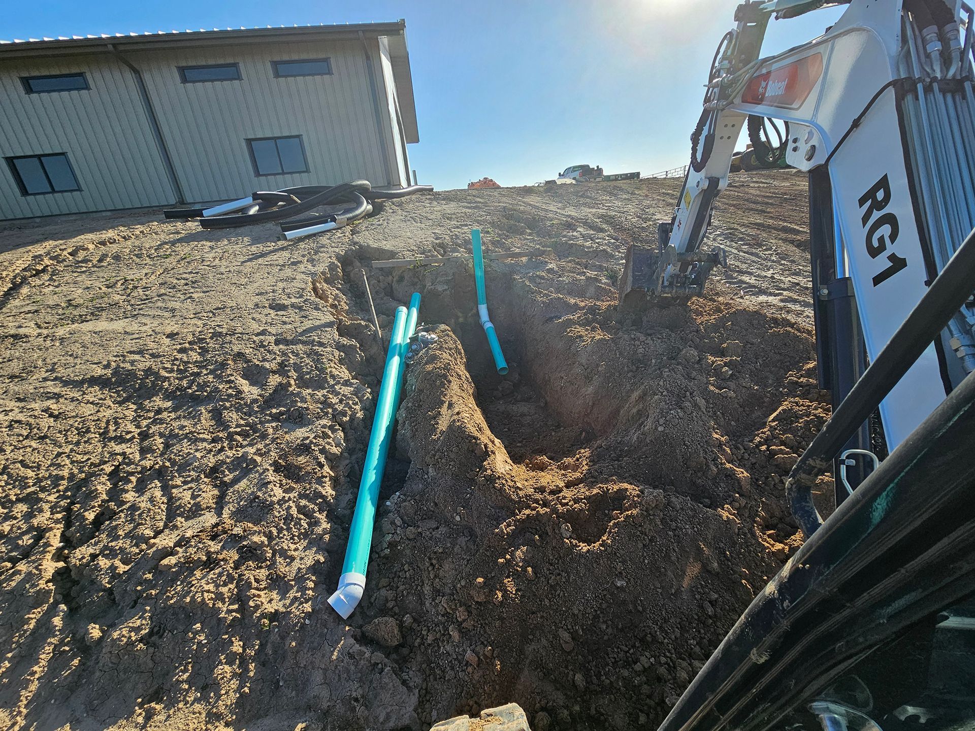 Excavation with a backhoe, revealing buried green pipes on a hillside; house in the background.