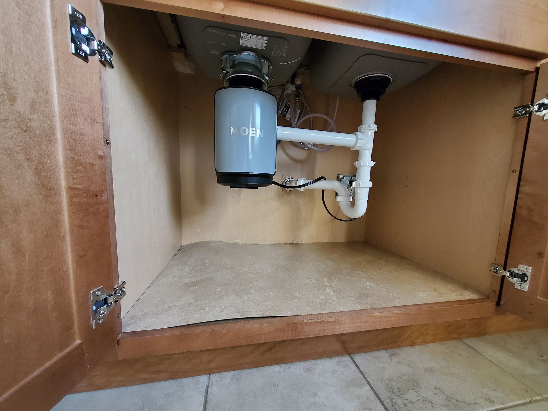 Kitchen cabinet open, showing sink, garbage disposal, and plumbing. Brown wood, tan interior, and white pipes.