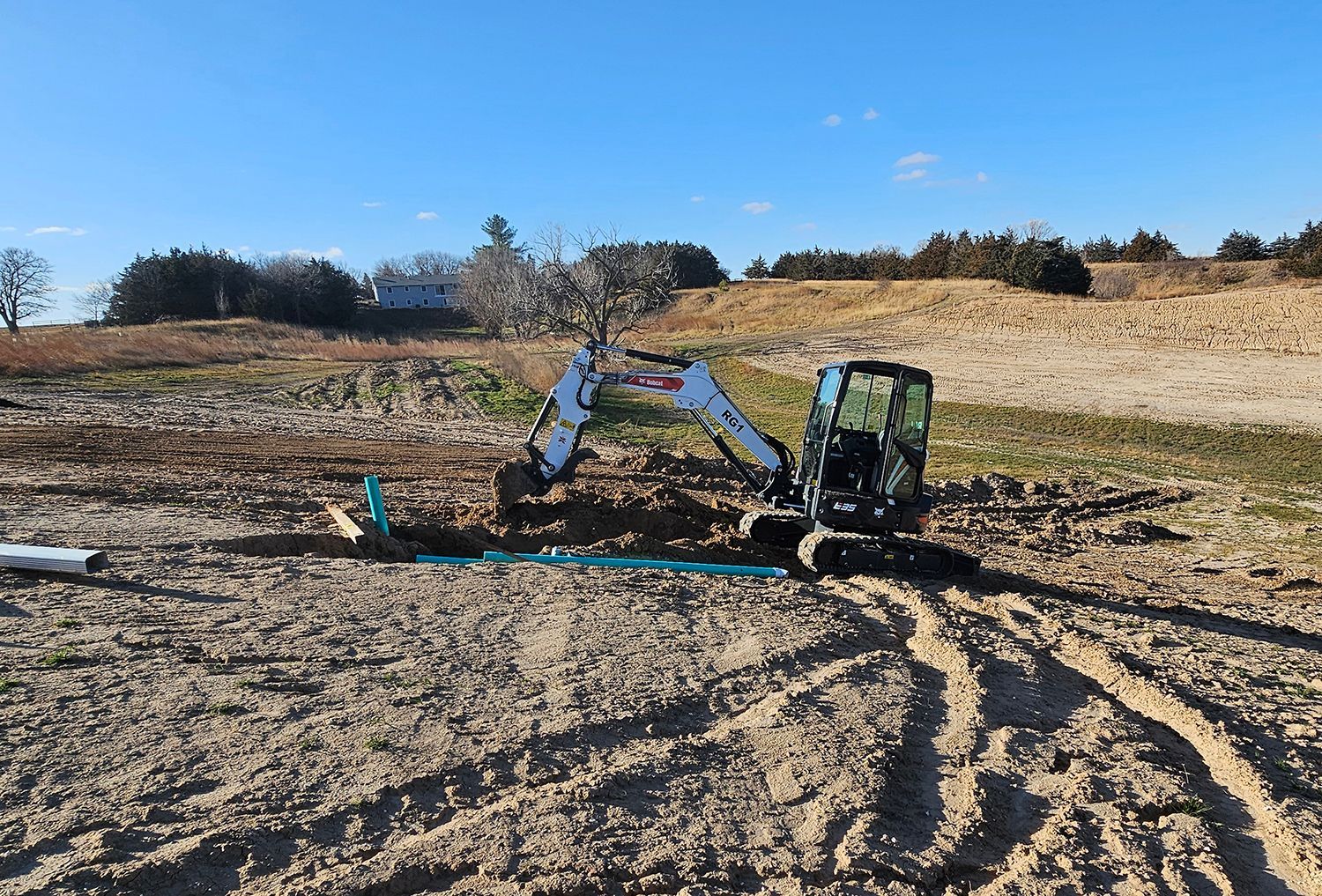 Mini excavator digging in a field, near blue pipes and a hill, under a clear sky.