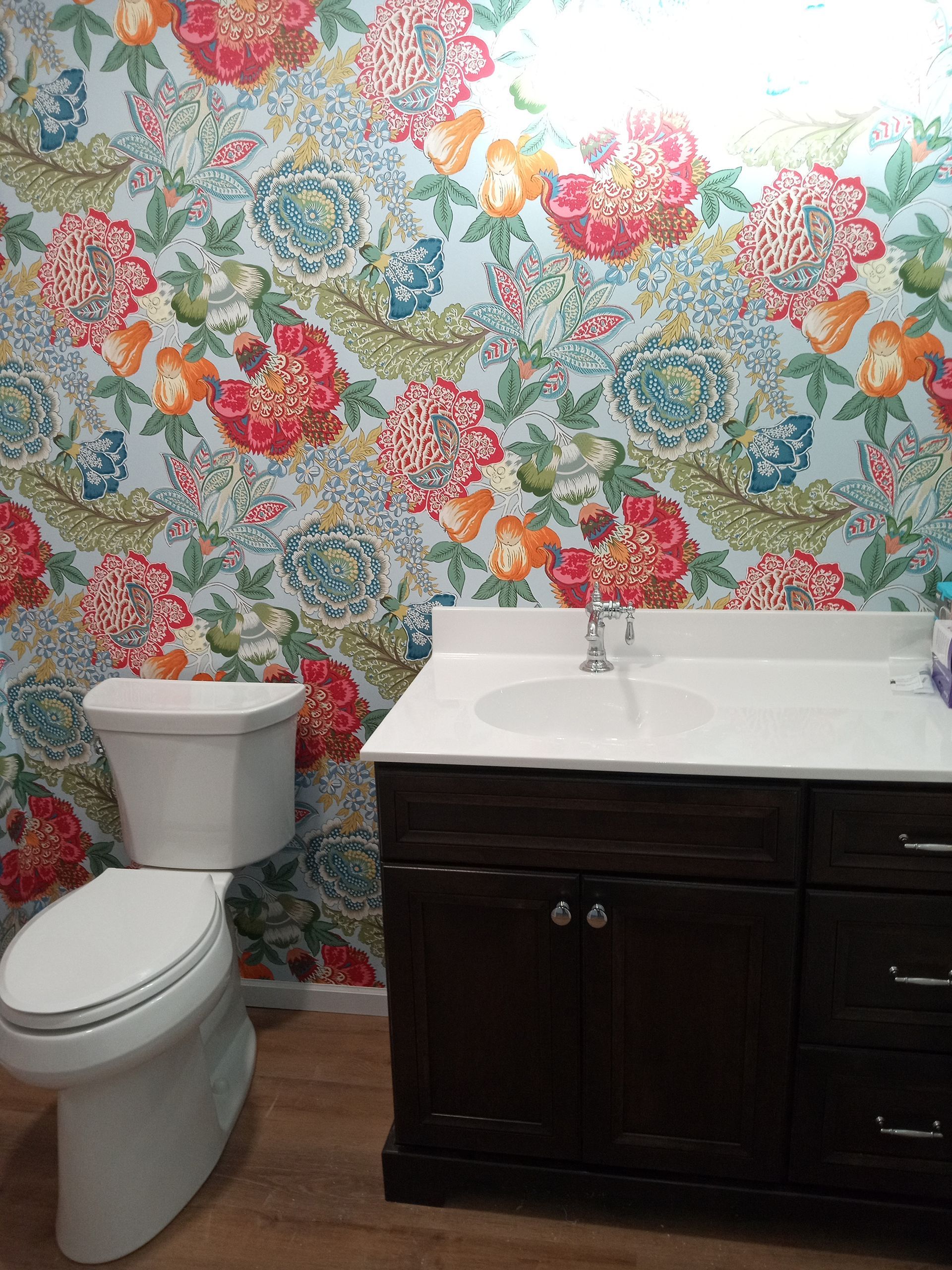 Bathroom with floral wallpaper, toilet, dark brown vanity, white sink, and wood-look floor.