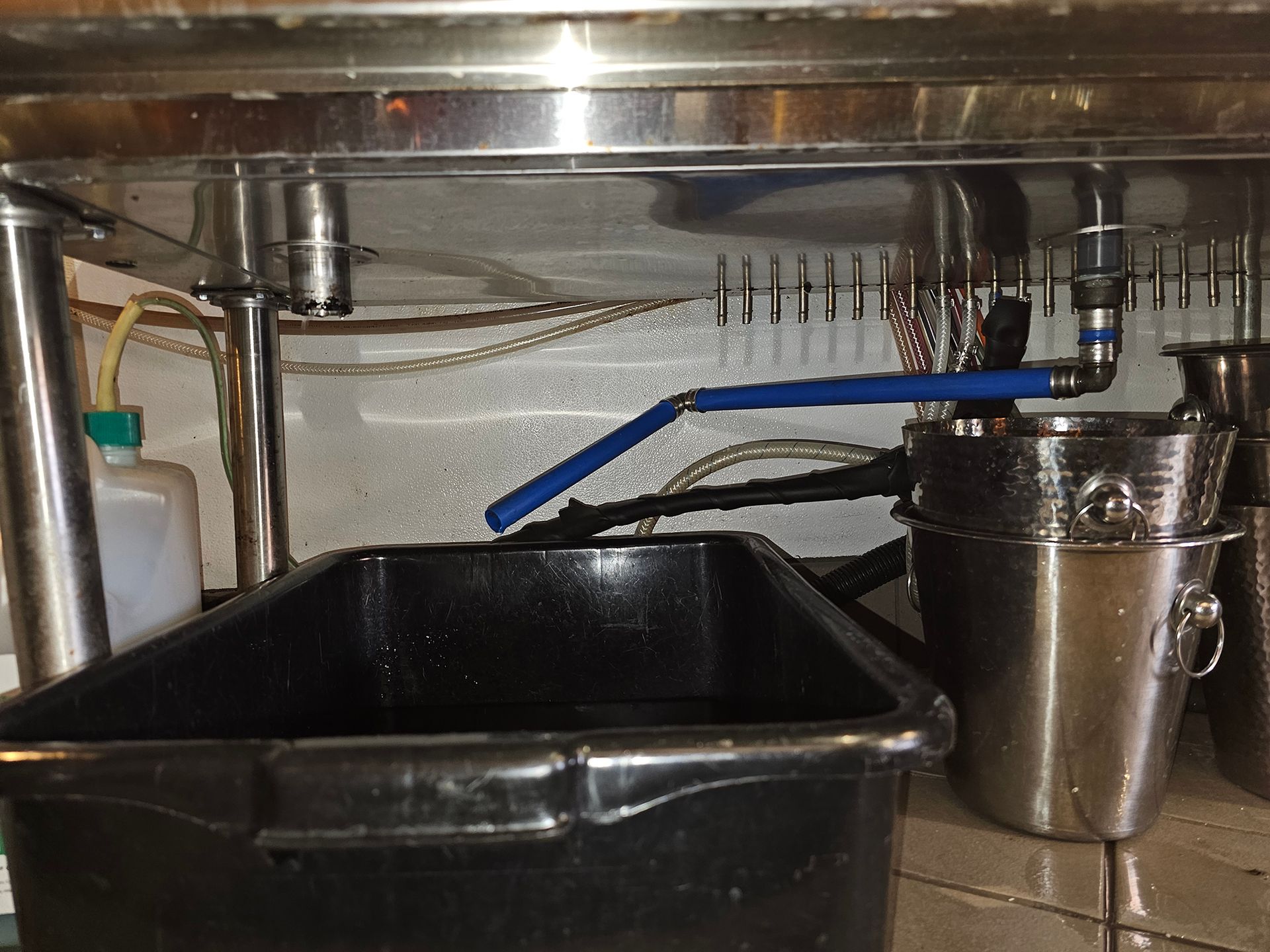 Underside of a stainless steel bar with a blue pipe draining into a black bin, with metal buckets on the right.