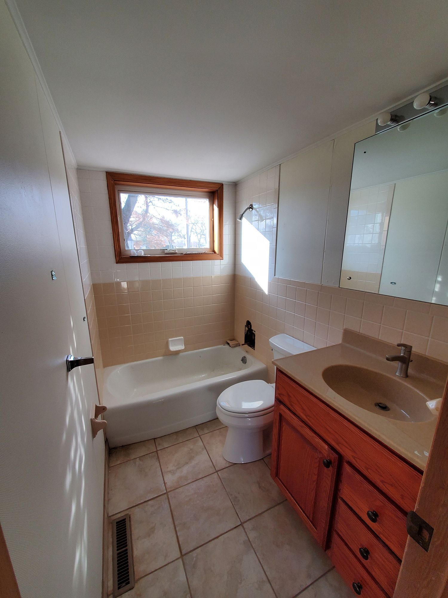 Bathroom with a bathtub, toilet, vanity, and window. Beige tile, wood trim, and a mirror.