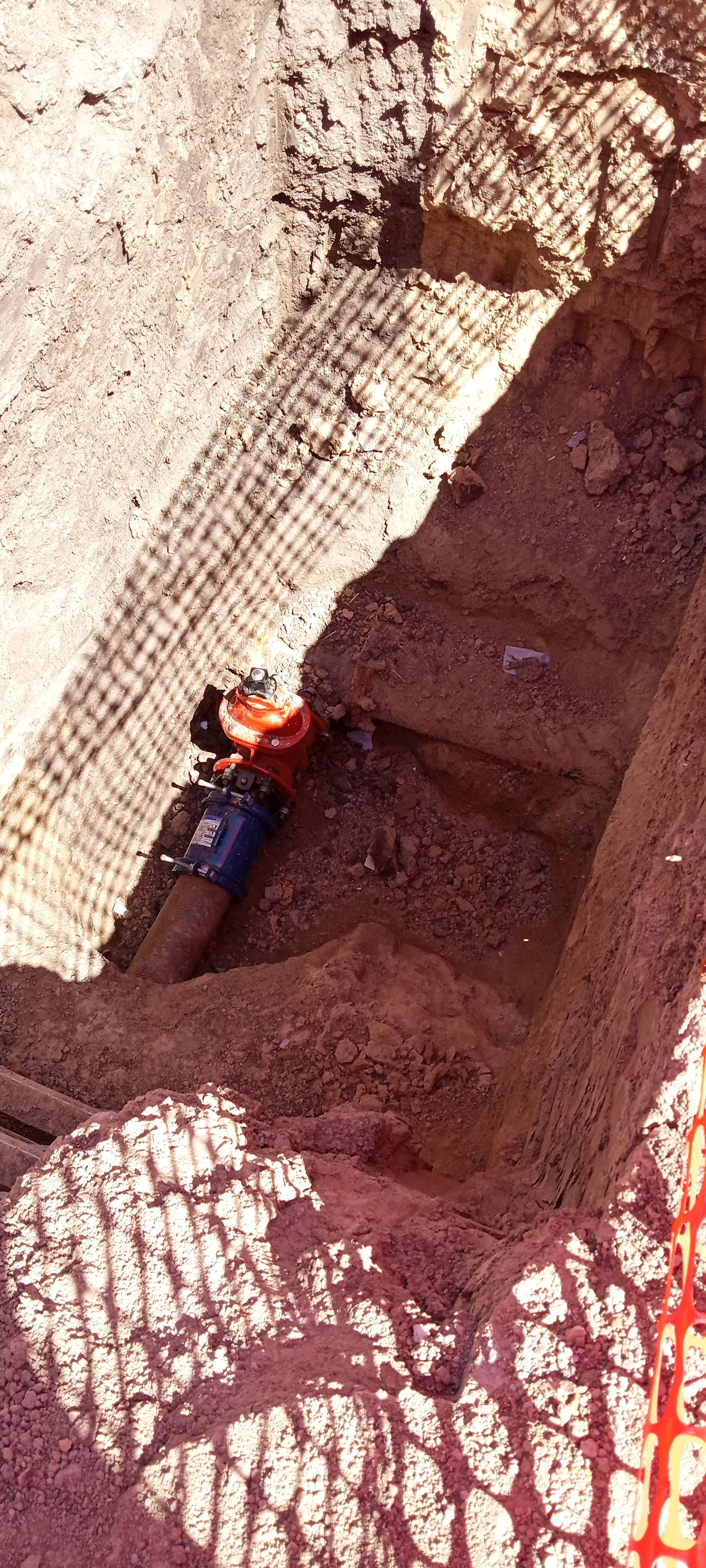 A water pipe connection in a trench is being worked on. Orange and blue pipe fixtures are present.