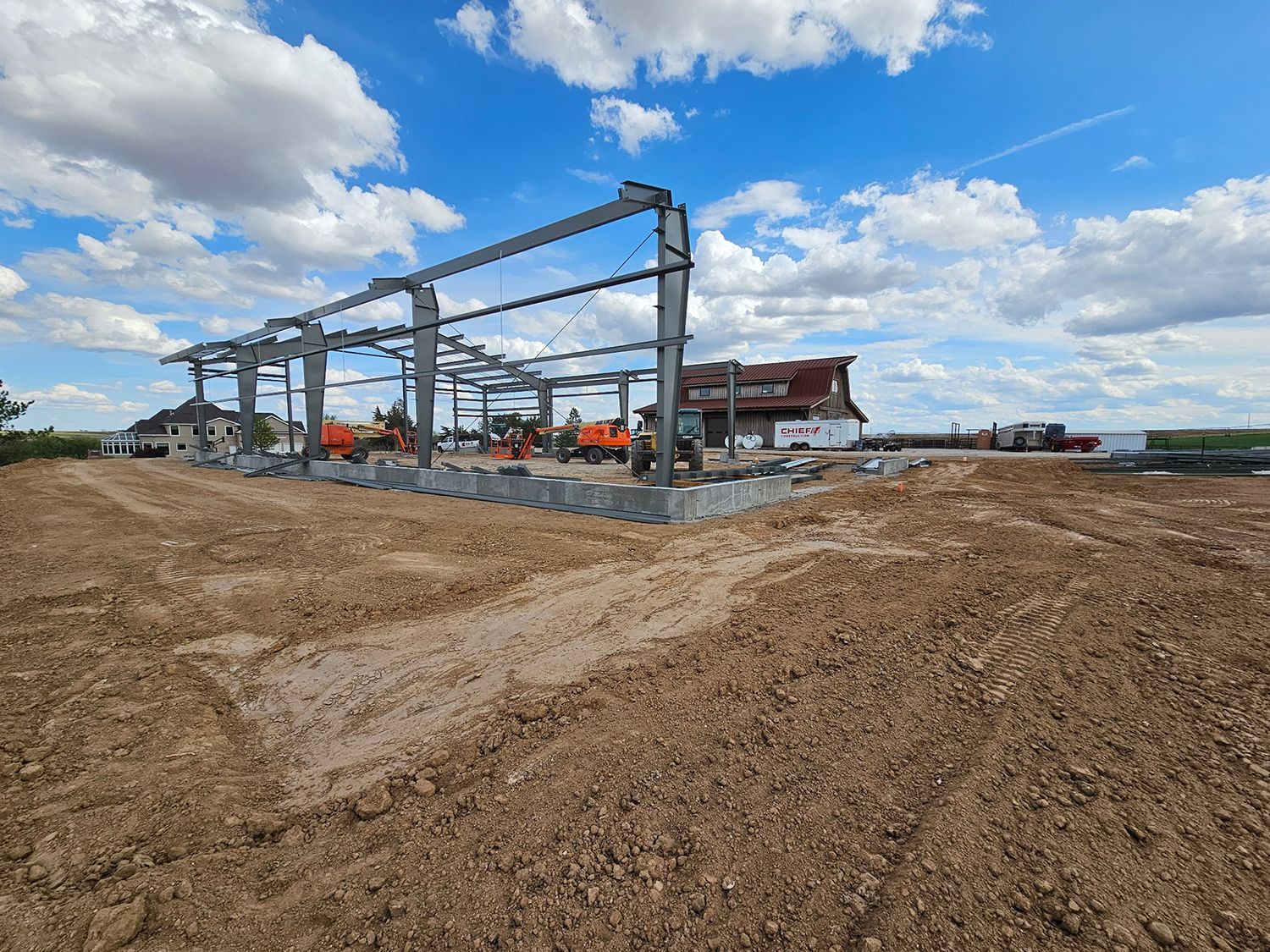 Steel framework of a building under construction on a dirt lot, cloudy blue sky in background.
