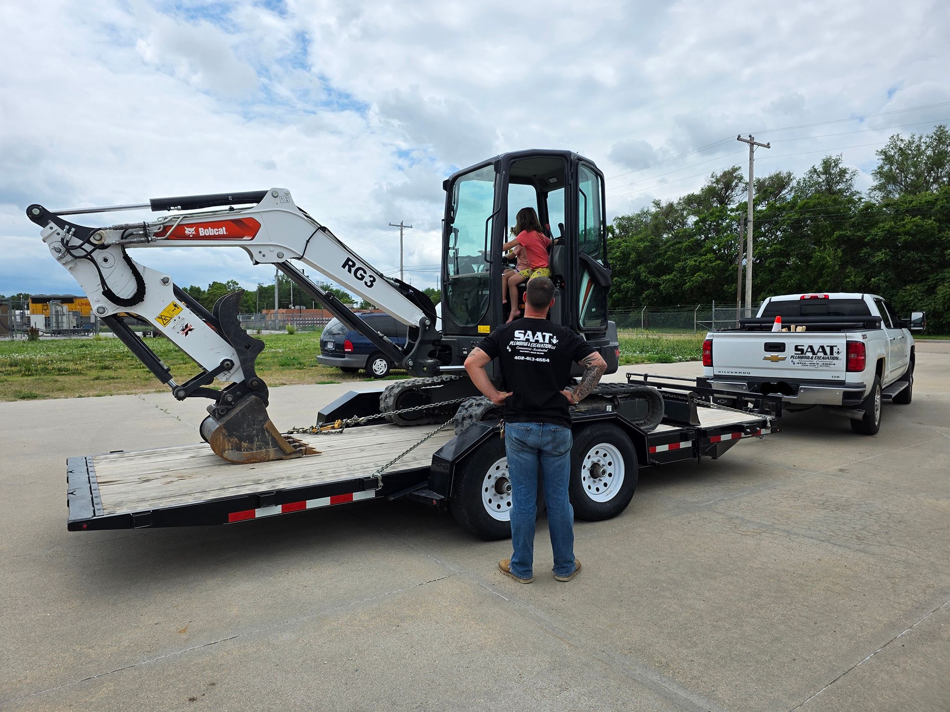 Man stands near a trailer with a Bobcat excavator, hitched to a truck, outdoors. Another person sits in the excavator.