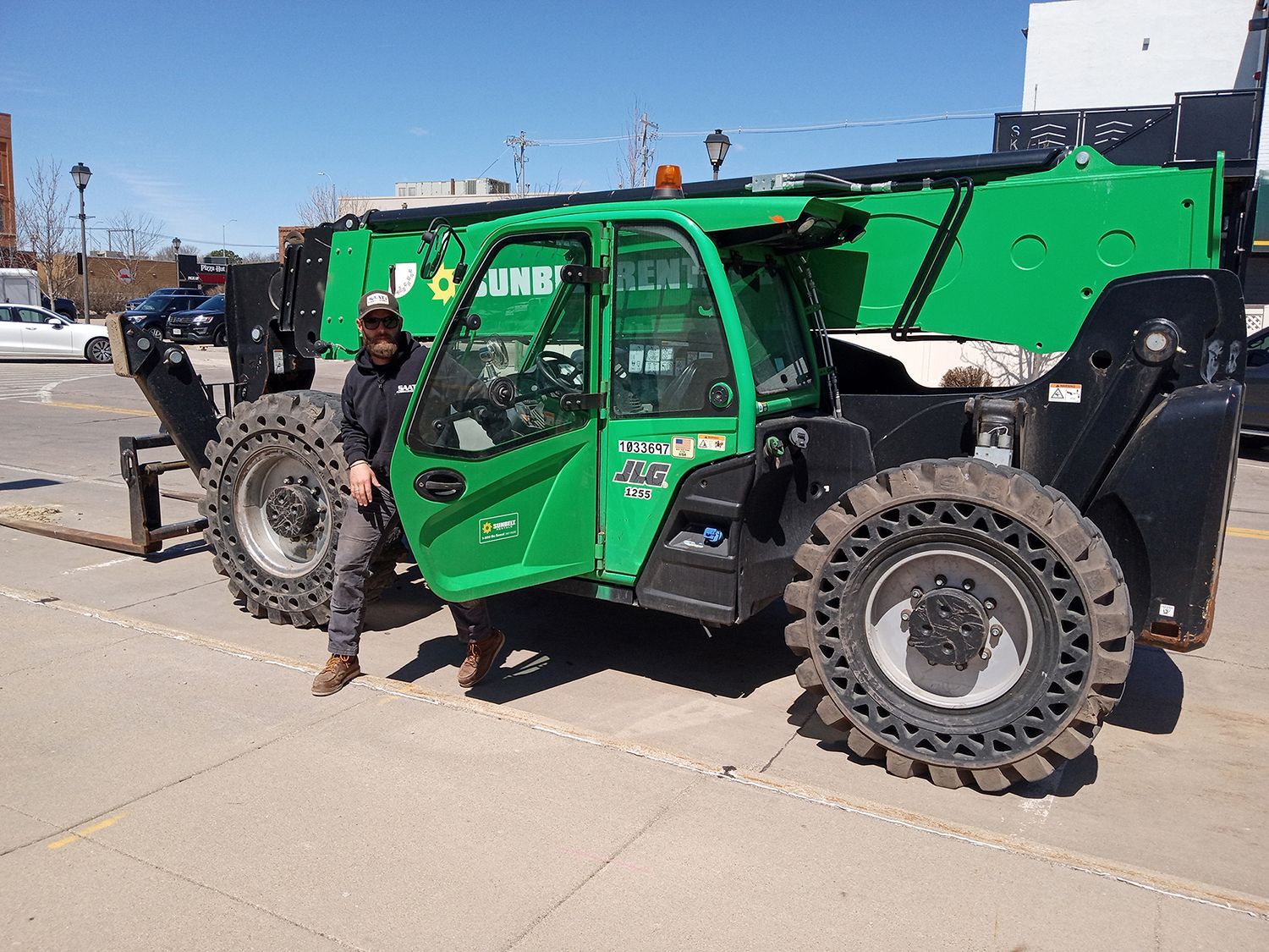 Man stands next to a large green telehandler. The machine is parked on pavement with cars in the background on a sunny day.