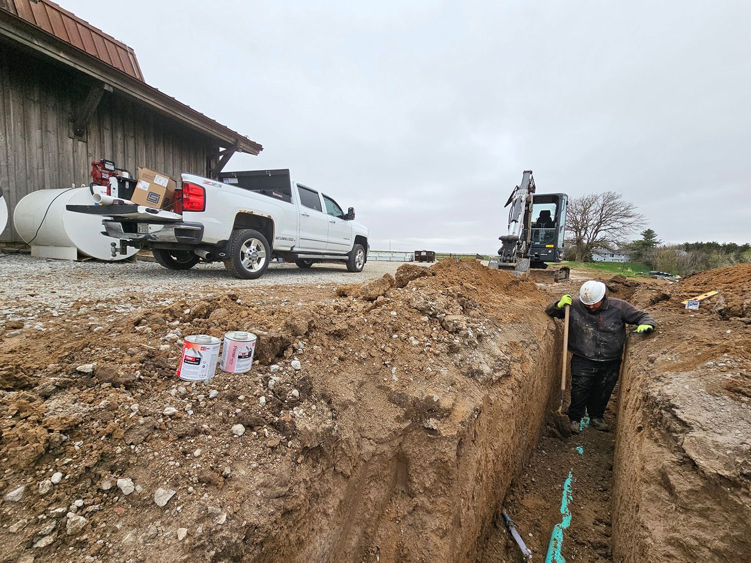 Man in trench working on pipes, truck and excavator nearby on a construction site.