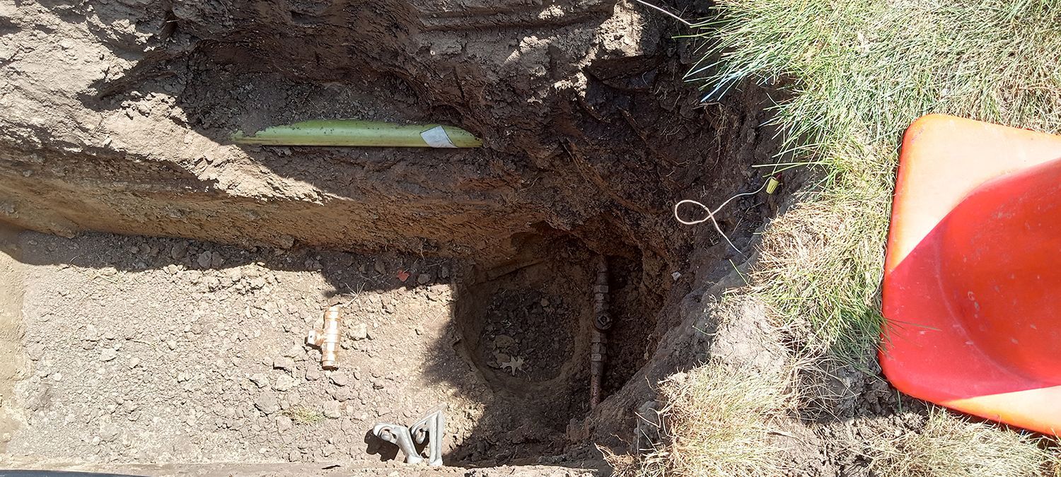A trench with a green pipe visible, next to grass and an orange cone.