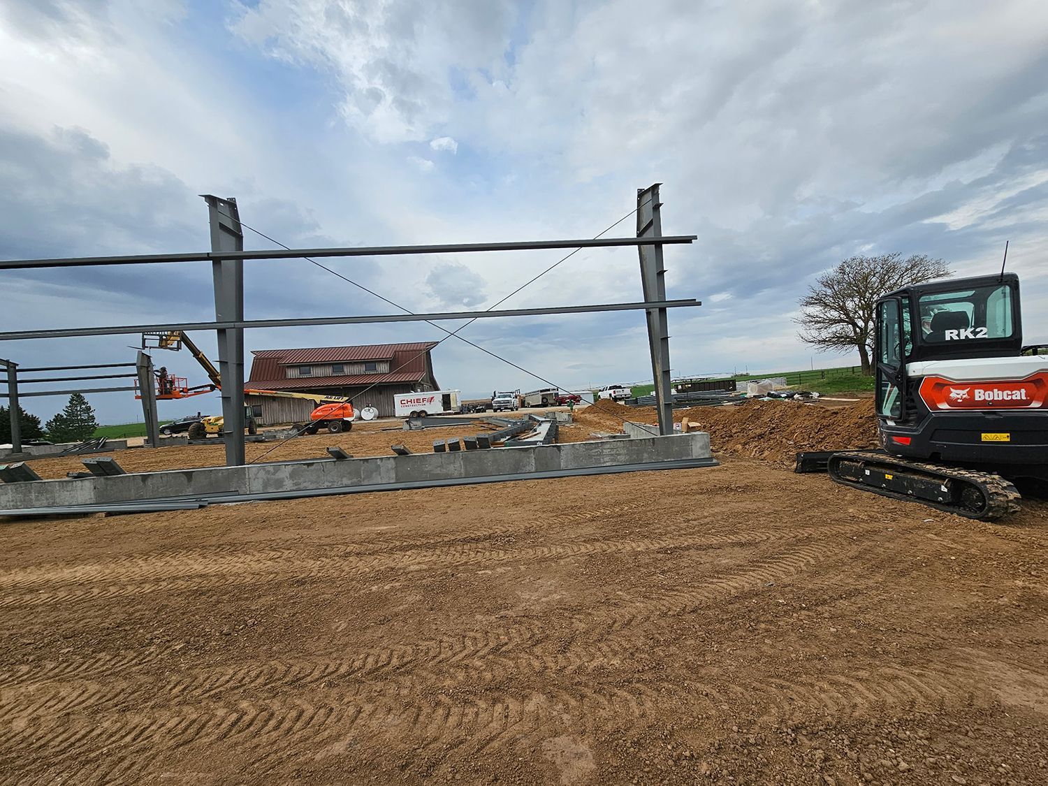 Construction site with steel frame, excavator, and leveled ground under a cloudy sky.