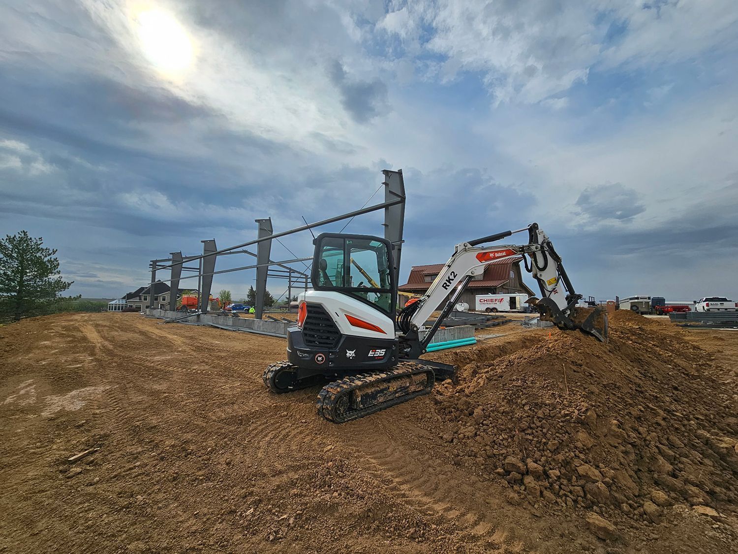 A small white excavator on a construction site with a partially built steel structure in the background. Cloudy sky.