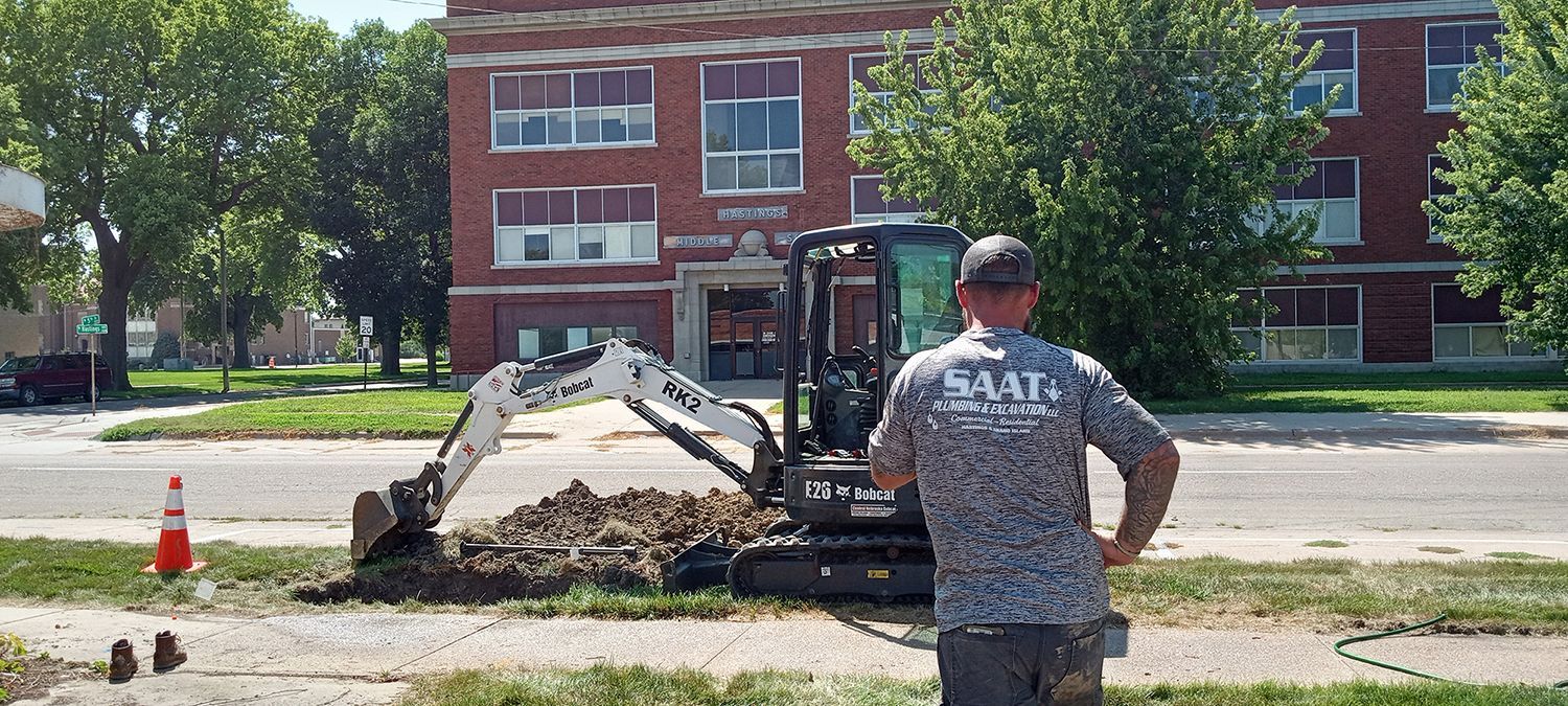 Man in a shirt with logo stands near a small excavator, working near a building with red brick facade.