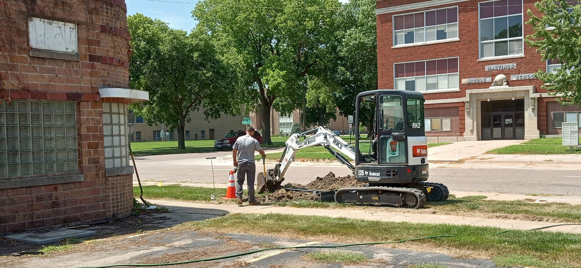 Construction worker using an excavator on a residential street in front of a brick building.