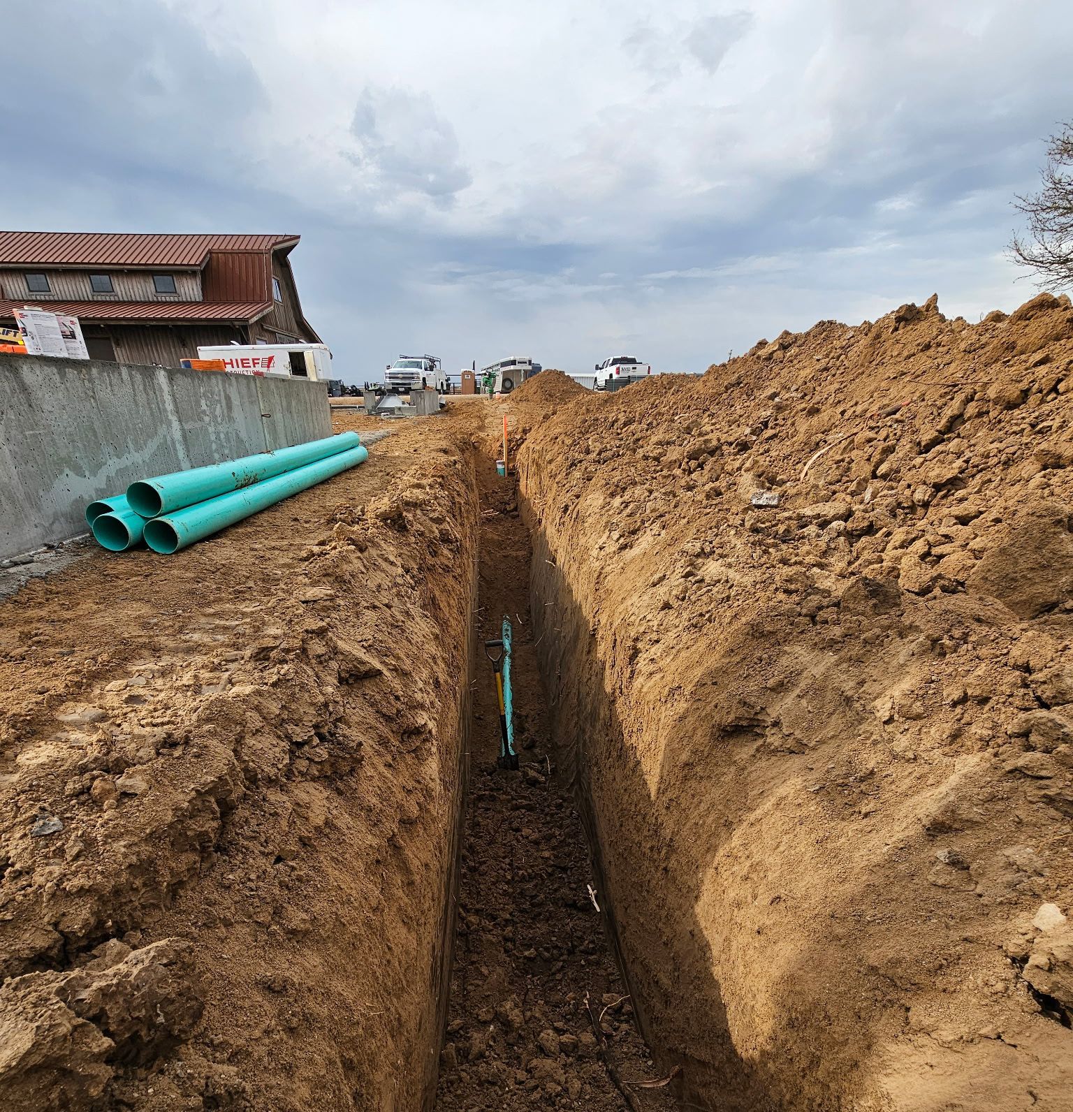 Trench dug in dirt with blue pipes beside it; construction site next to a building under cloudy sky.
