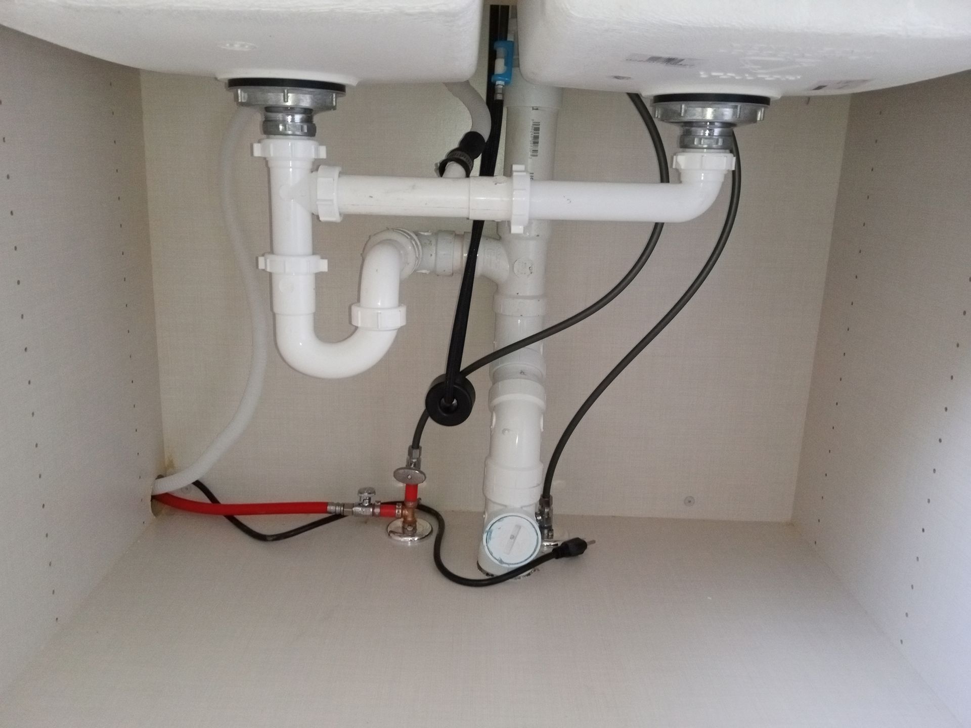Under kitchen sink: white plumbing pipes, U-bend, drain pipes, black wires, red water line, and shut-off valve.