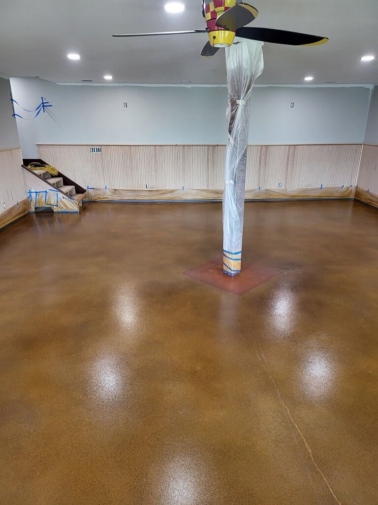 Brown stained concrete floor, a central pole wrapped in plastic, and a ceiling fan in a room.