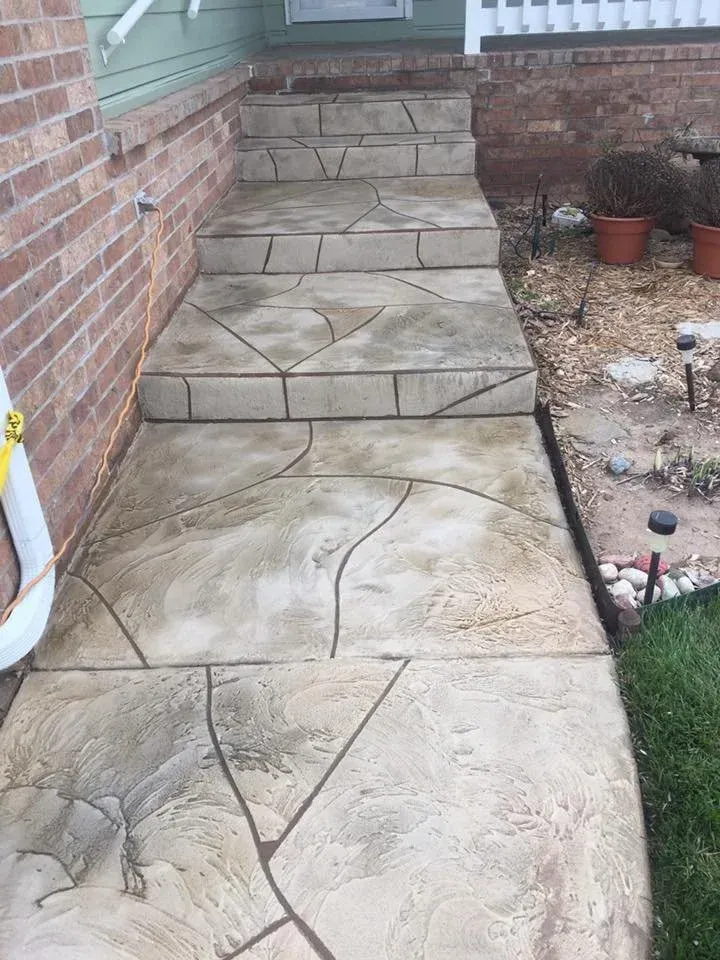 Concrete walkway with stone pattern leads up to brick home's entrance, bordered by grass and garden.
