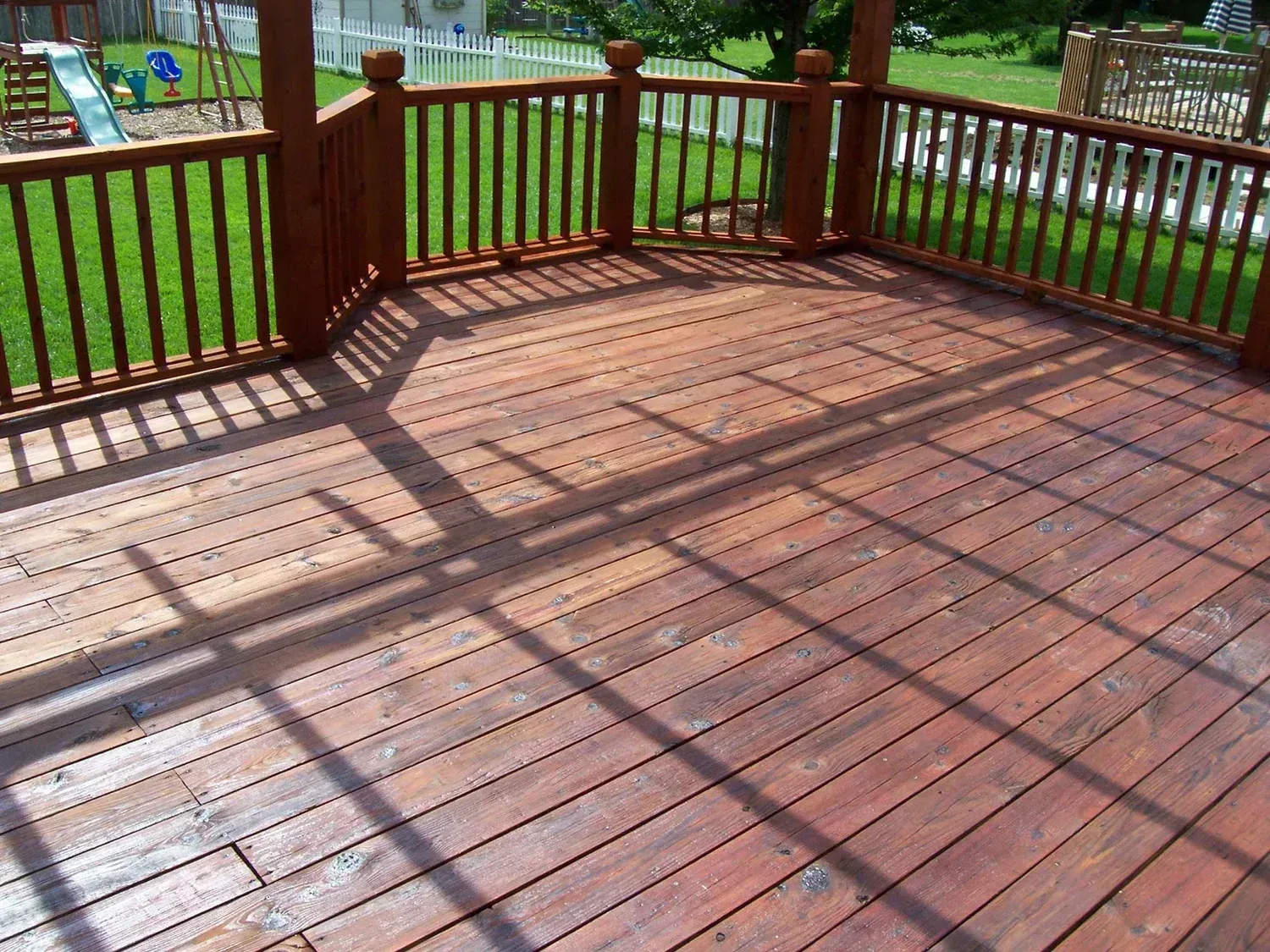 Wooden deck with railings and pergola casting shadows on red-stained planks; overlooking a grassy yard.