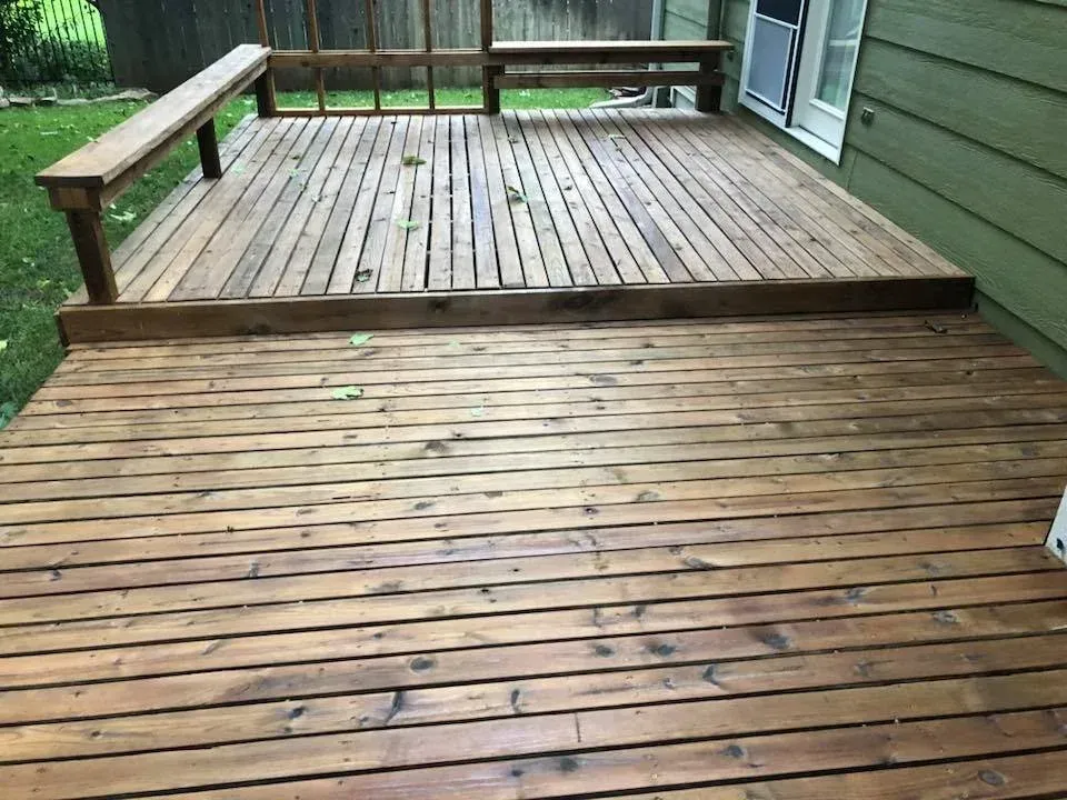 Wooden deck with bench and railing, attached to a green house, with grassy yard in the background.