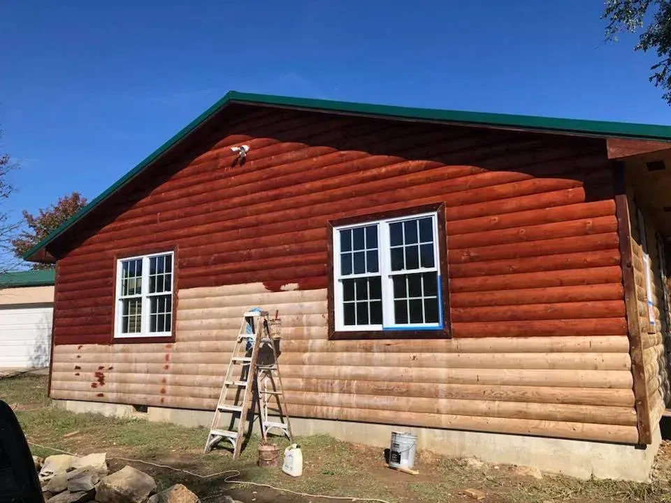 Side view of a cabin with wooden siding, partially painted red. A ladder is in front of the unpainted section.