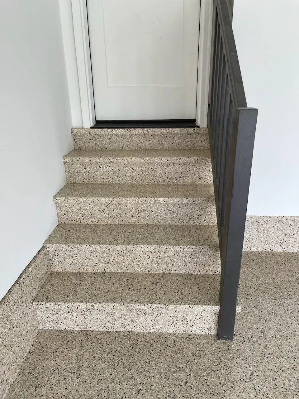 Staircase with speckled flooring leading to a white door. A dark gray railing is to the right. White walls.