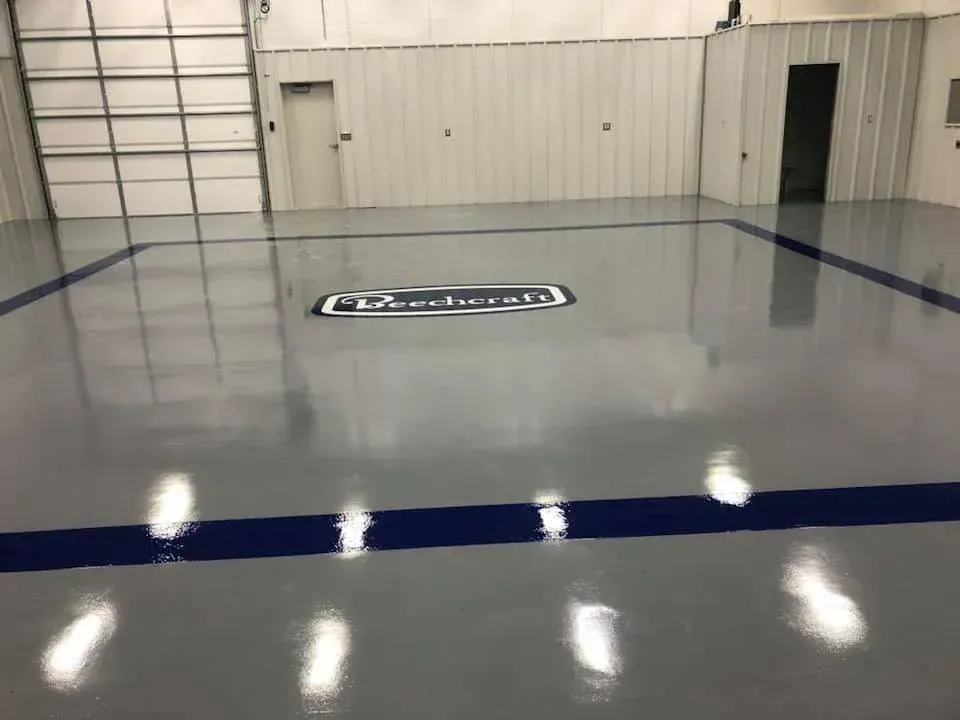Gray epoxy floor in a warehouse with blue border and logo in the middle, overhead lights reflected.