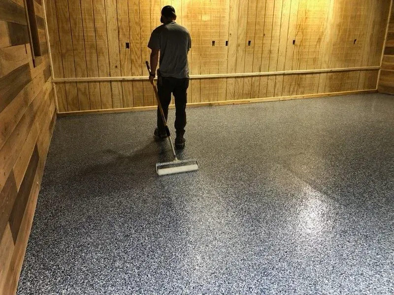 Person applying epoxy coating to a speckled gray floor in a room with wood paneling.