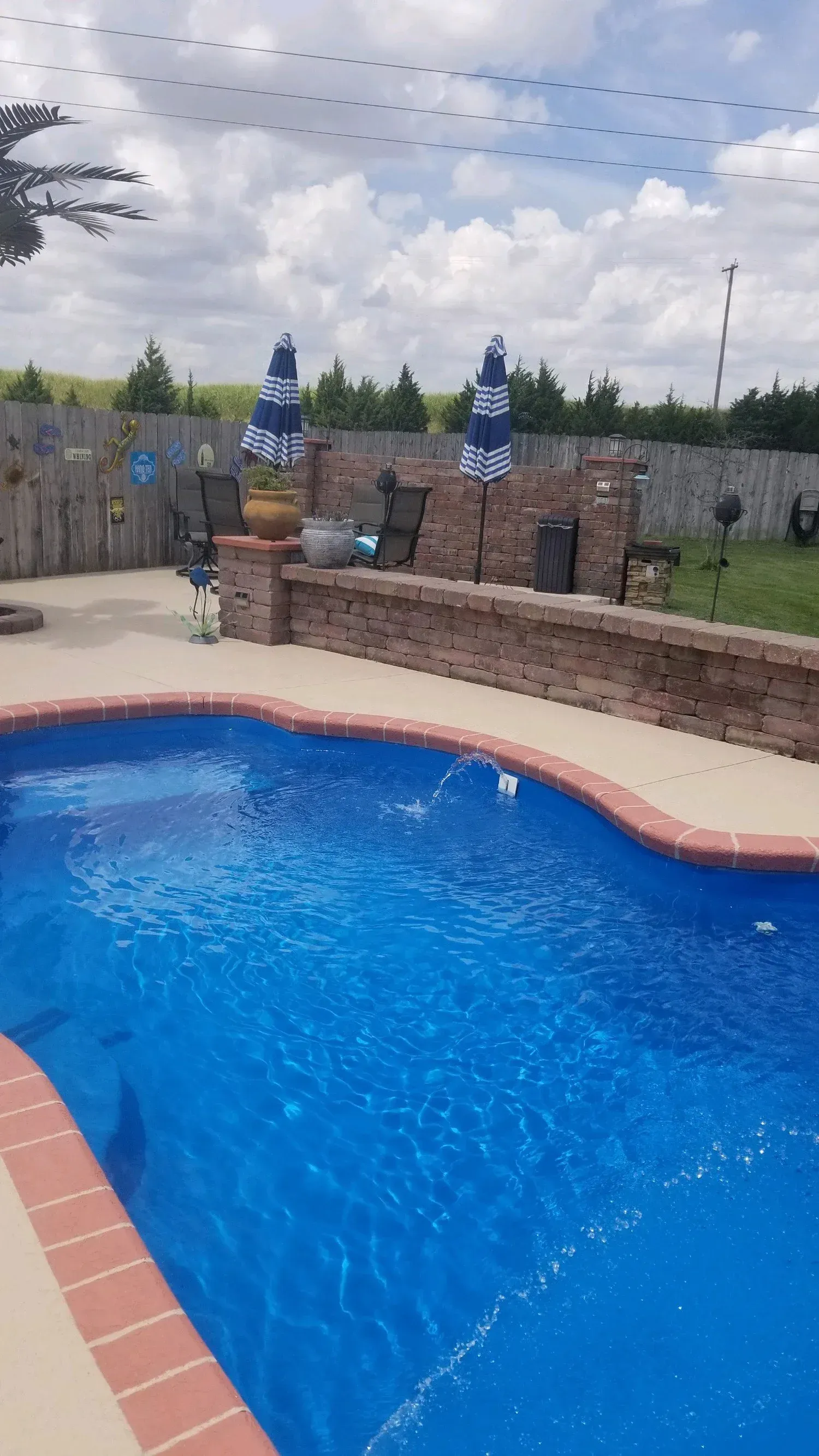Swimming pool with brick border, patio, and blue and white striped umbrellas. Cloudy sky.