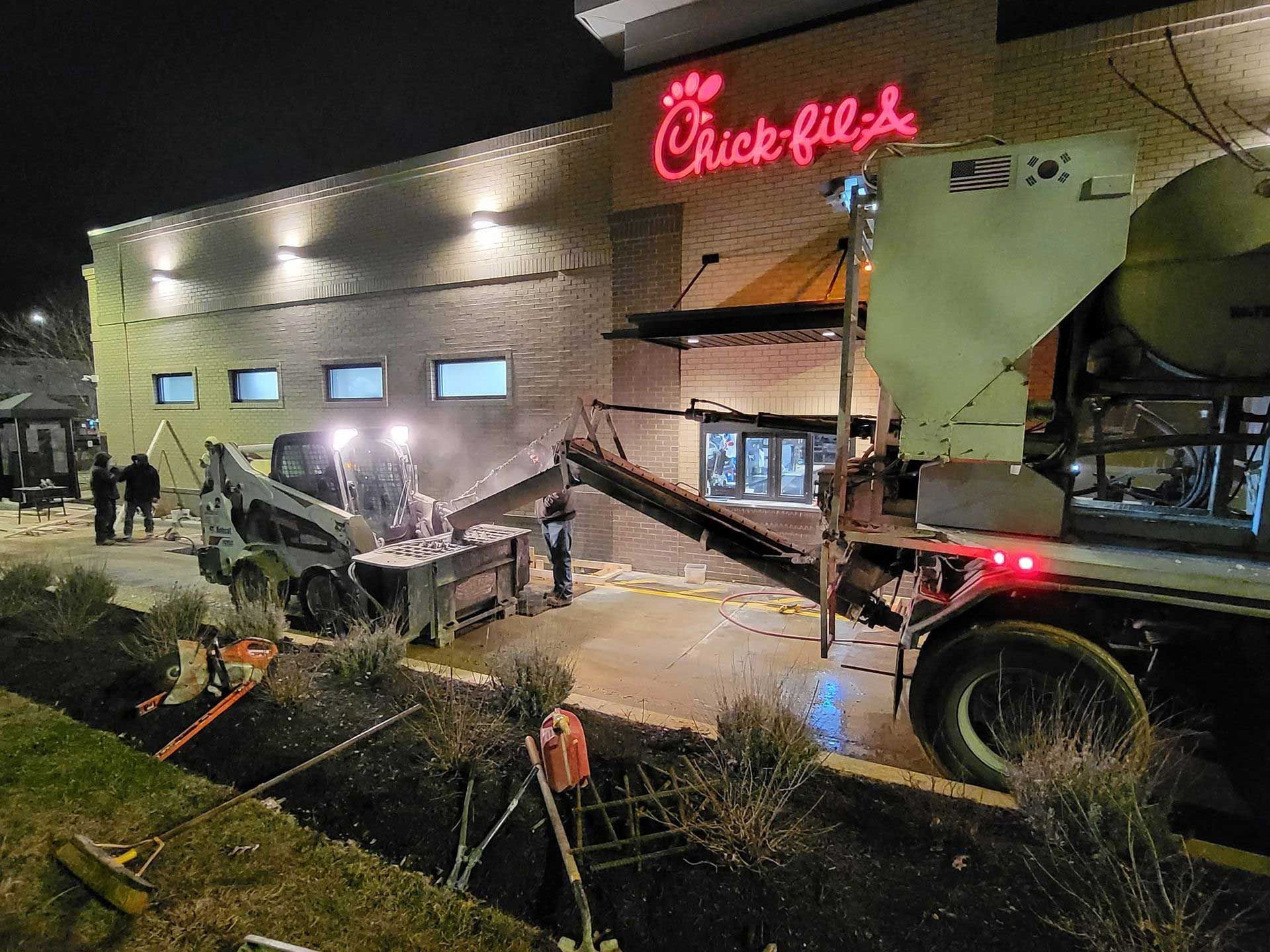 Construction at Chick-fil-A at night: a truck pouring gravel into a Bobcat while workers observe.