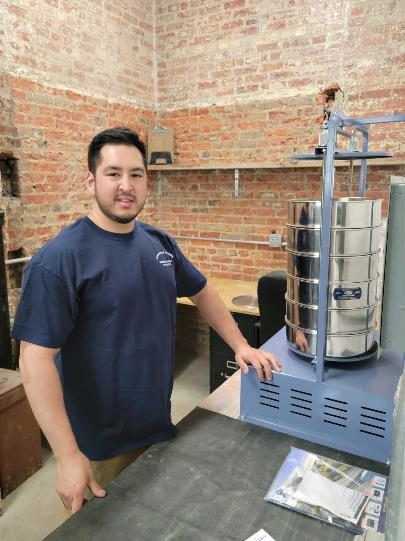 Man in blue shirt stands by metal kiln in brick room.