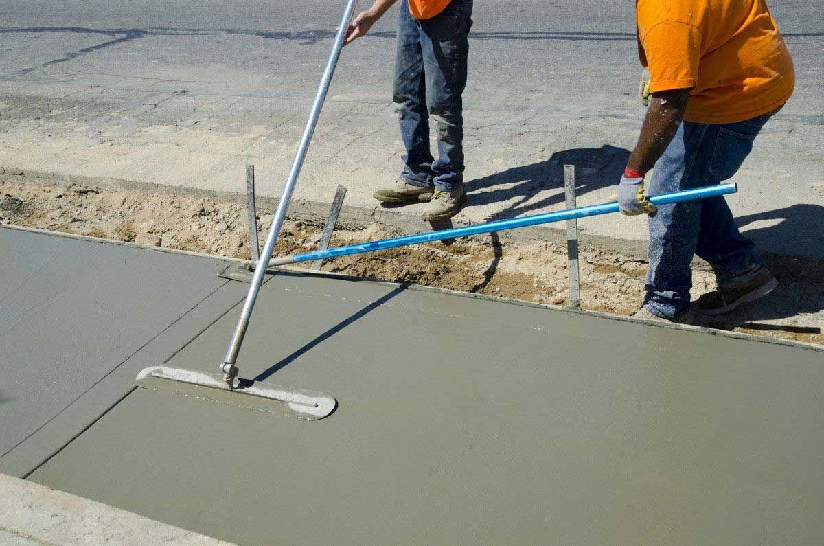 Workers smoothing wet concrete with a long tool, creating a sidewalk outdoors.