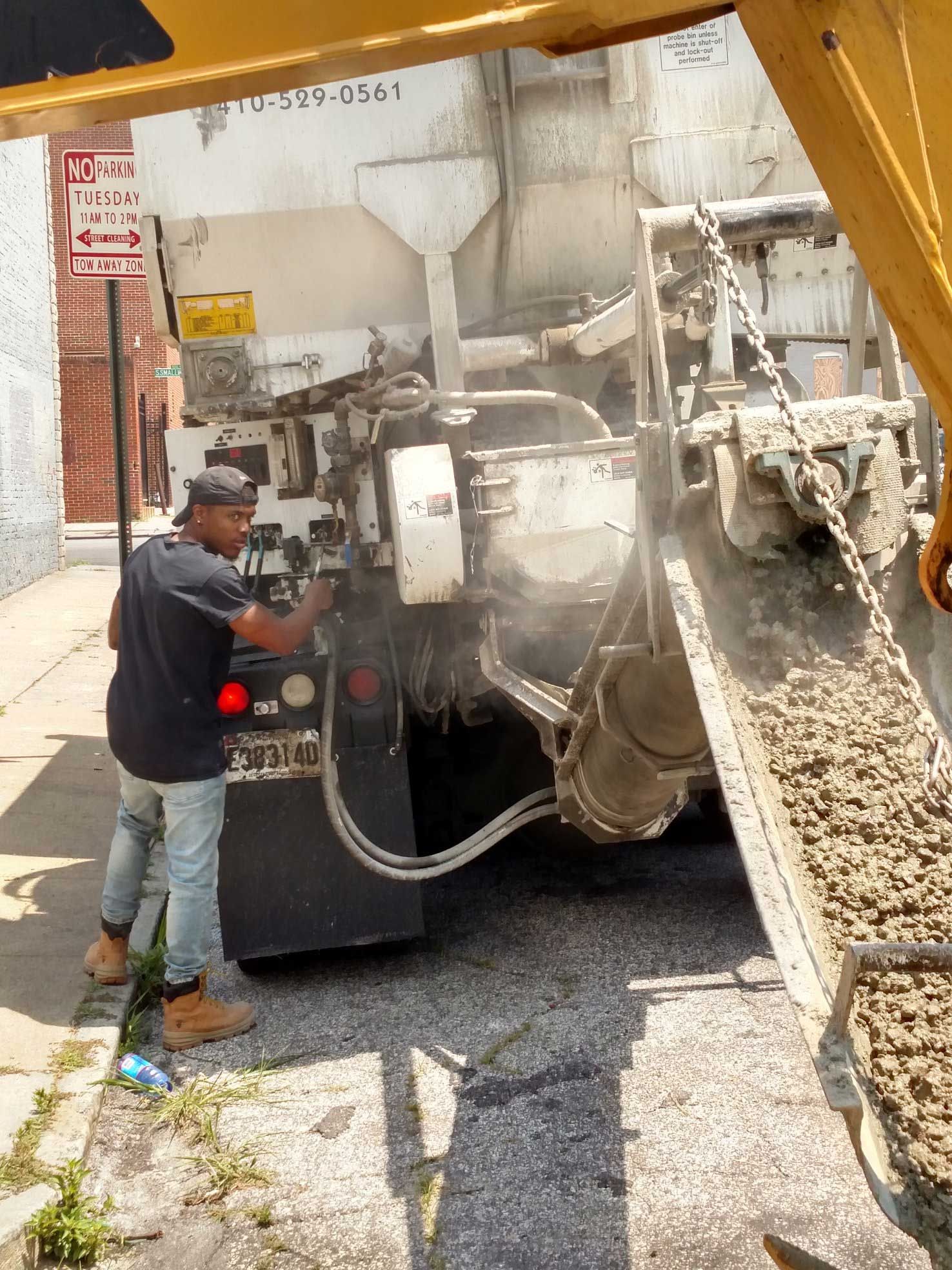 A person operates concrete mixer truck, pouring concrete on a street.