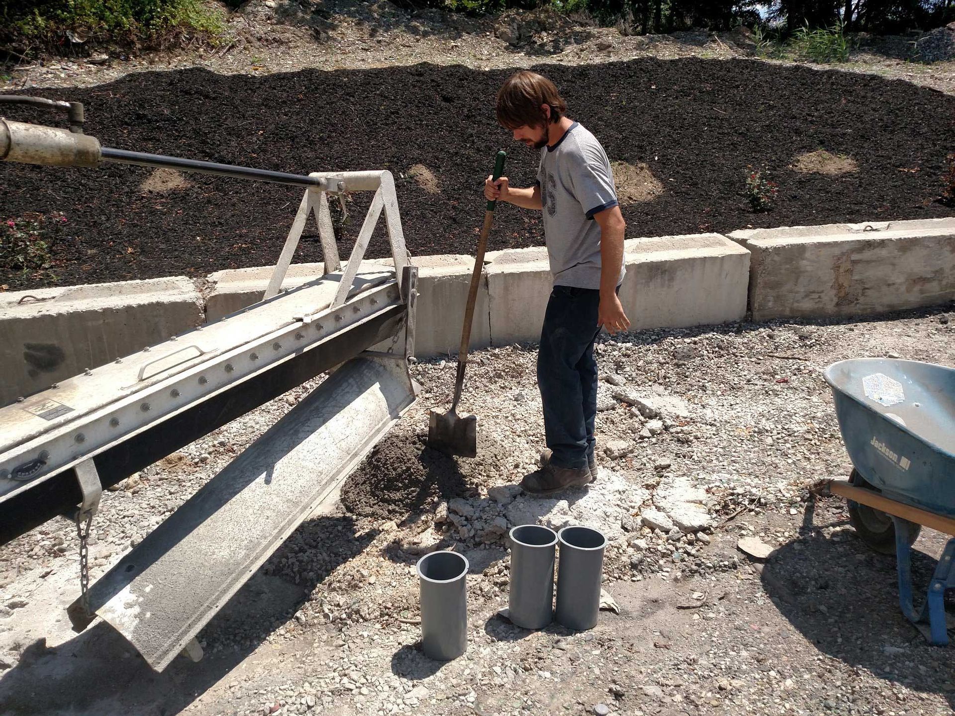 Man using a long tool to fill three gray cylinders with dirt samples, near concrete blocks and a retaining wall.