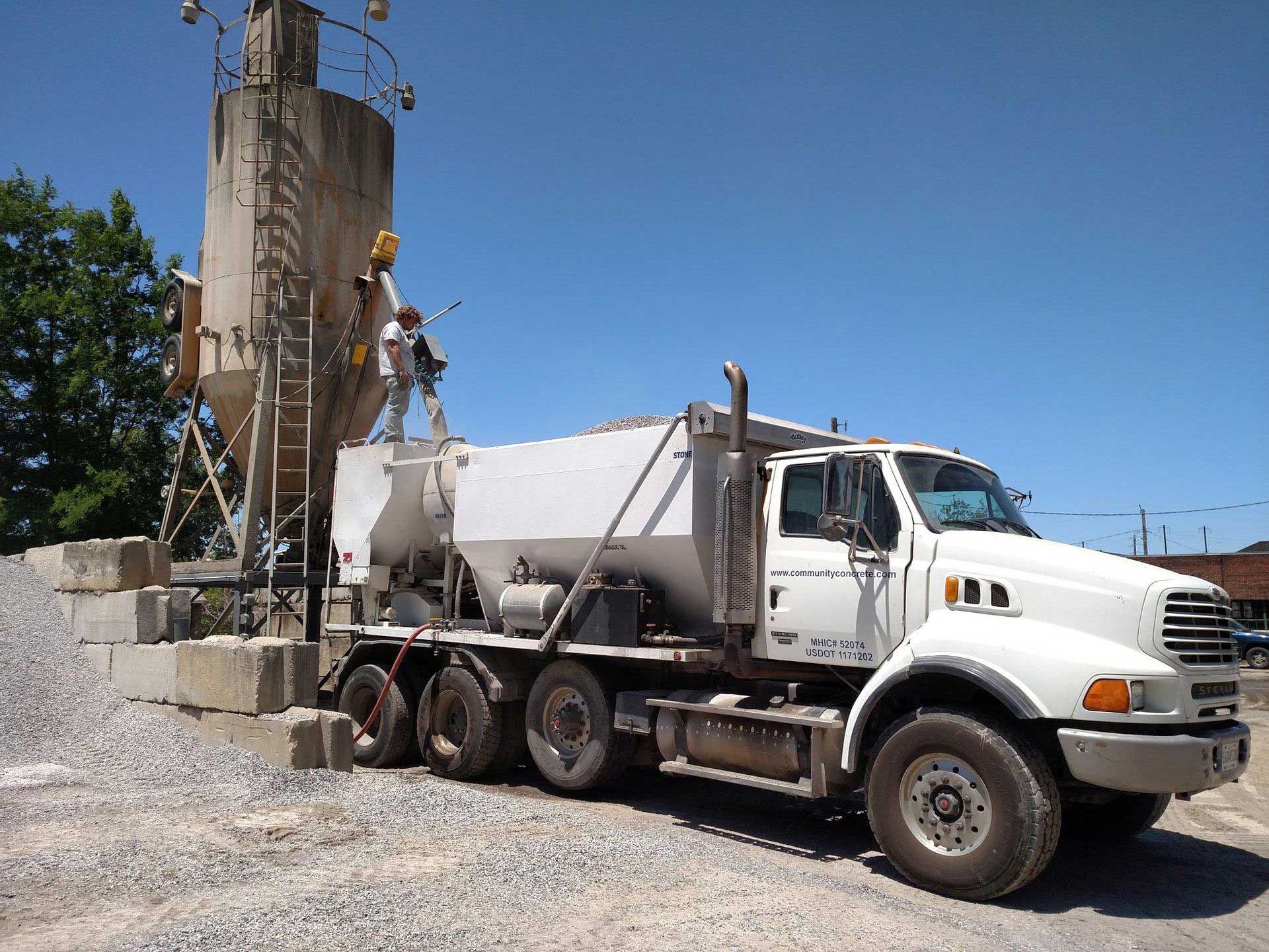 A white cement truck at a silo, workers filling the truck, gravel and concrete blocks sit nearby, blue sky.