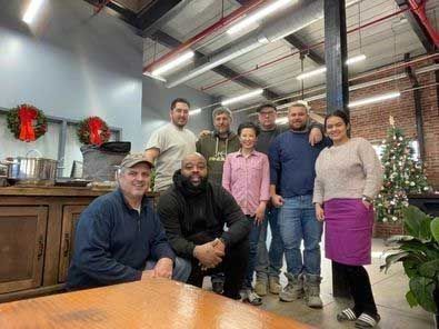 Group of diverse people smile at the camera indoors, near holiday decorations, including a Christmas tree.