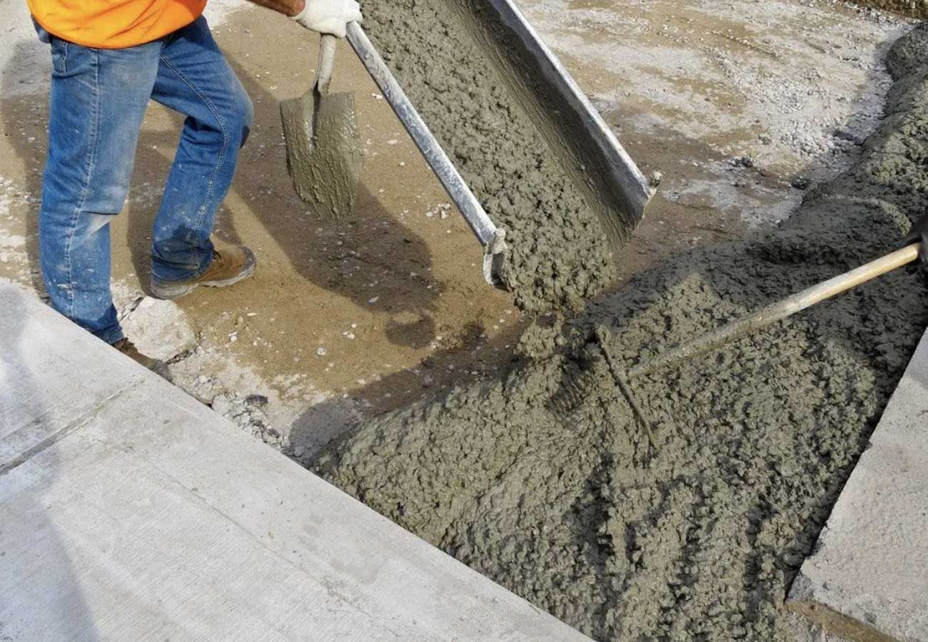 A construction worker pours concrete into a form, wearing gloves and jeans.