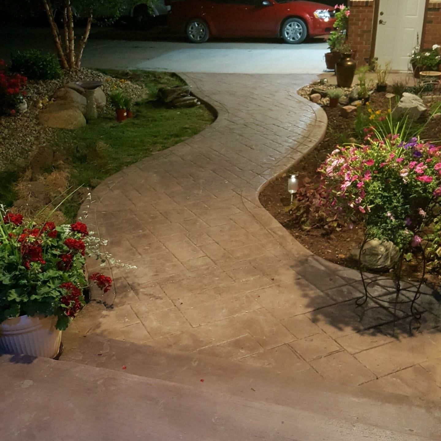 Curving concrete path with brick pattern, flowers, and landscaping leading to a house entrance; car parked nearby.
