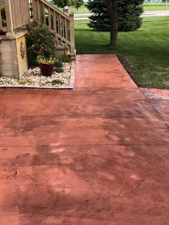 Stamped concrete walkway in reddish-brown color, leading from wooden porch steps to green lawn.