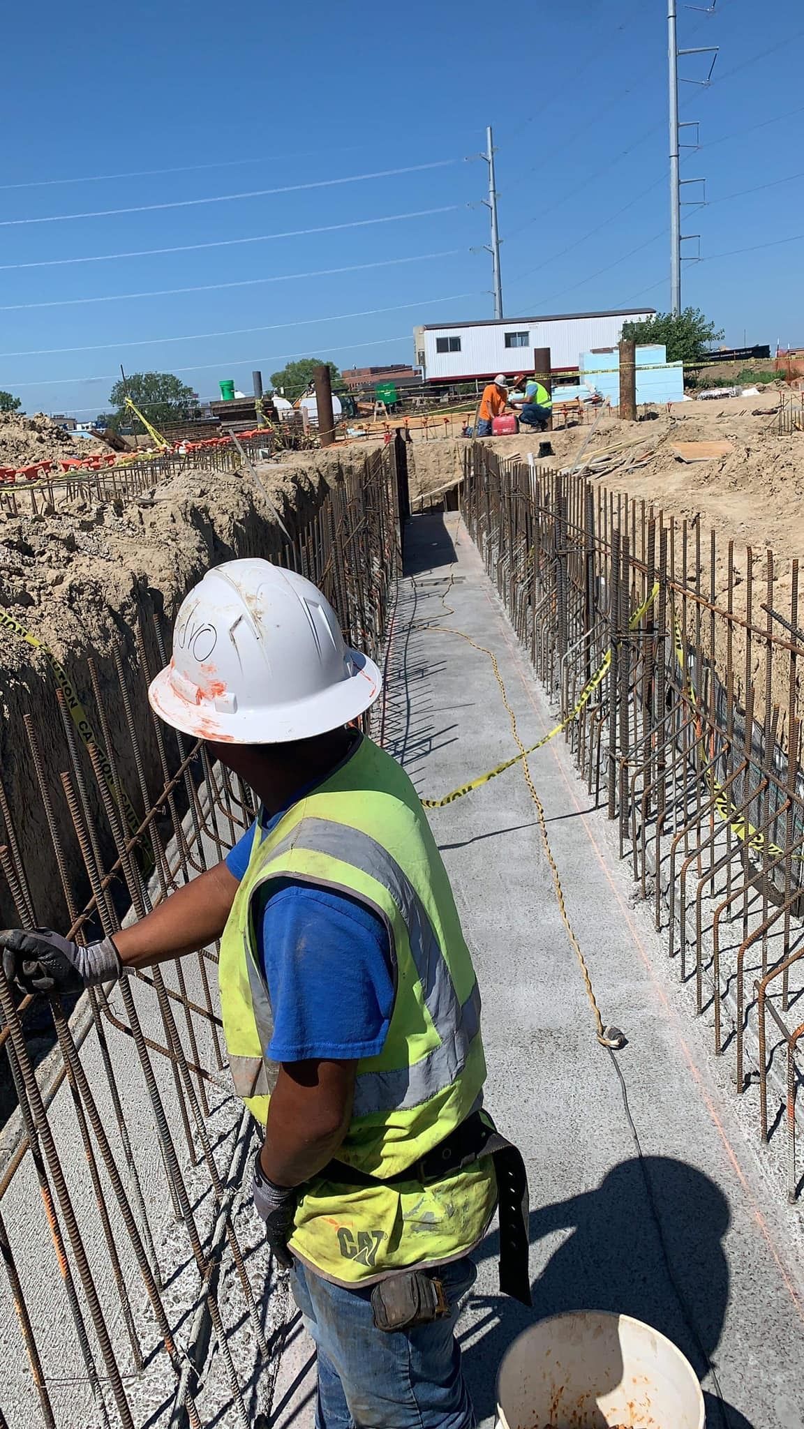Construction worker inspecting rebar in a concrete trench, wearing a hard hat and safety vest.