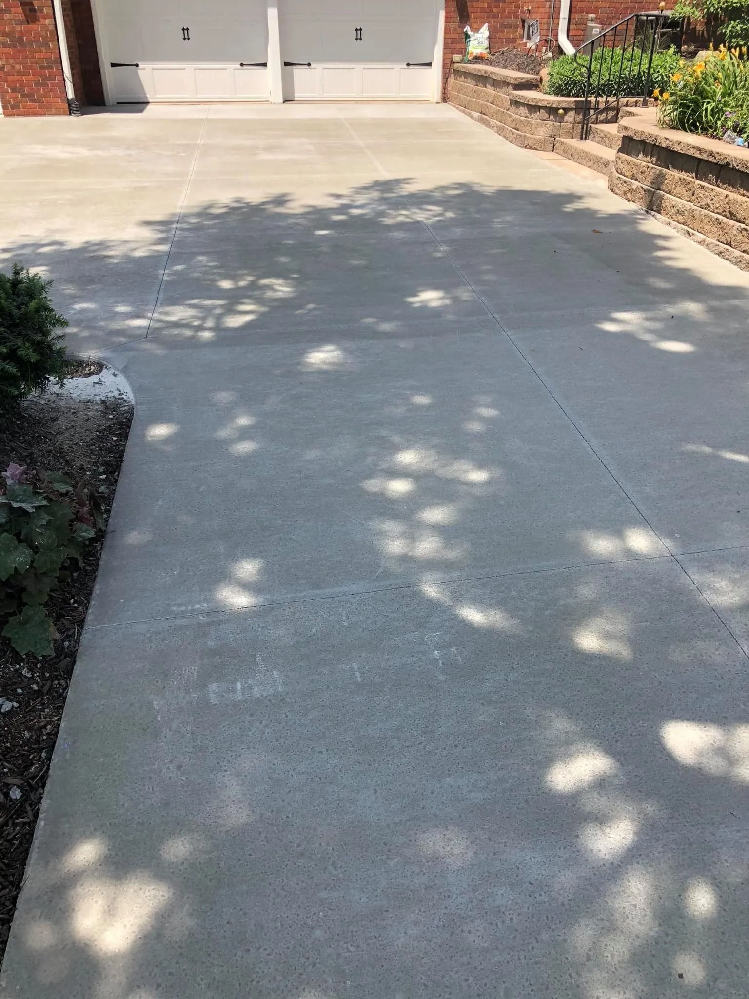 Driveway leading to a garage, with tree shadows. Edge has a flowerbed and brick retaining wall.
