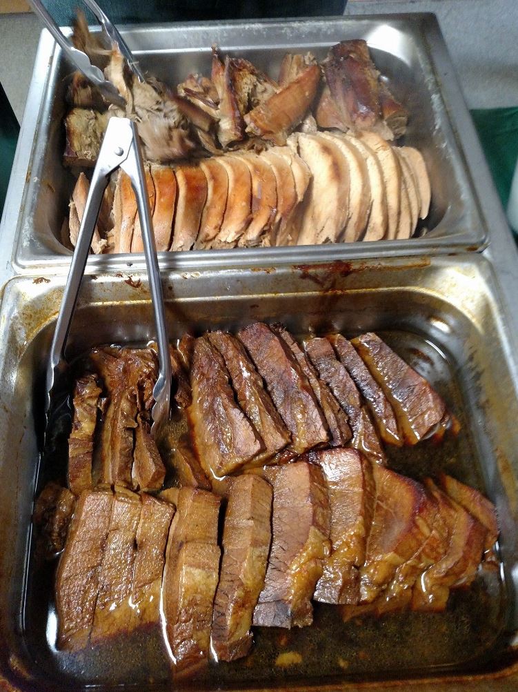 Two trays of meat are sitting on a table with tongs.
