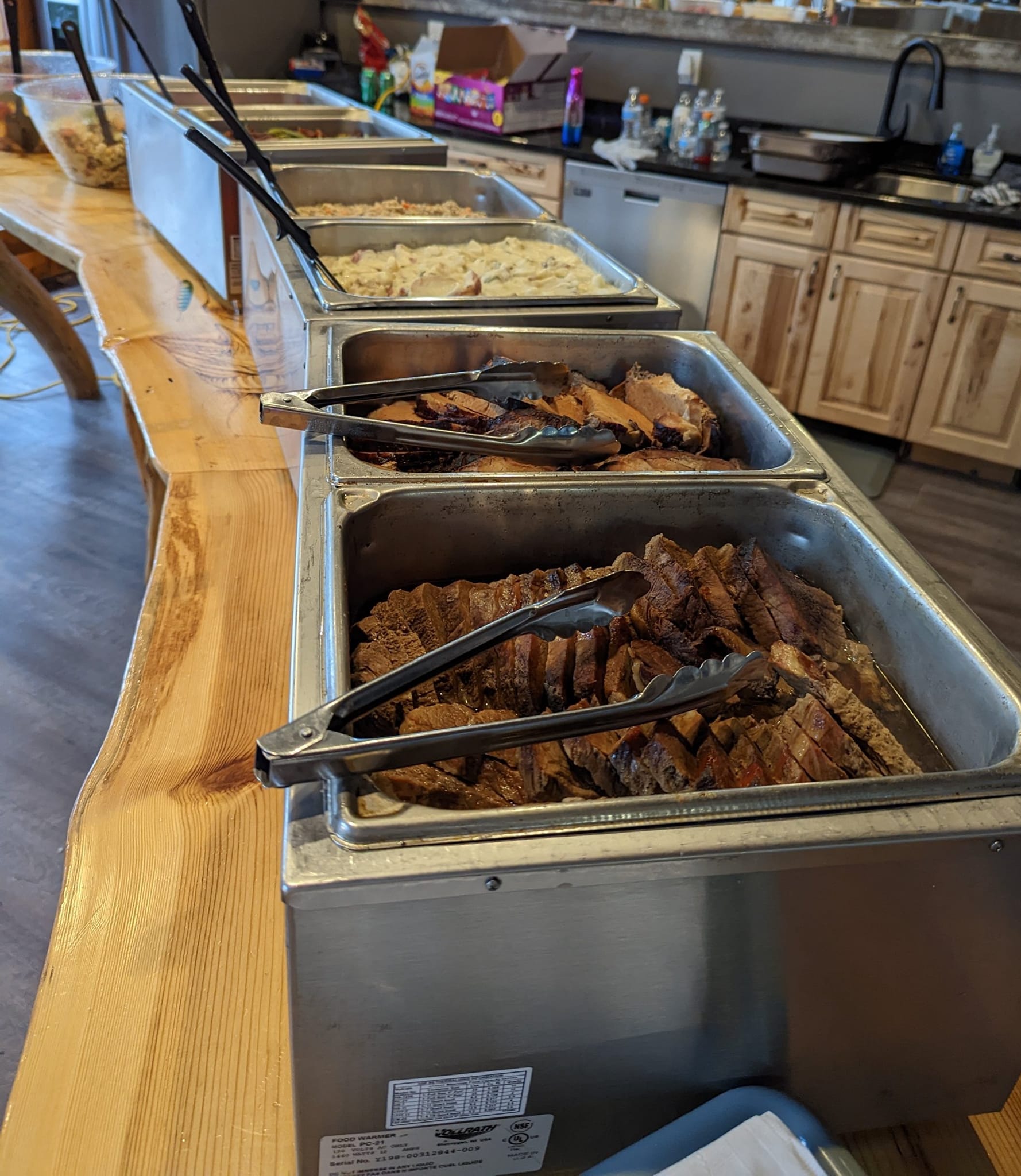 A buffet line with a variety of food and tongs on a wooden table.