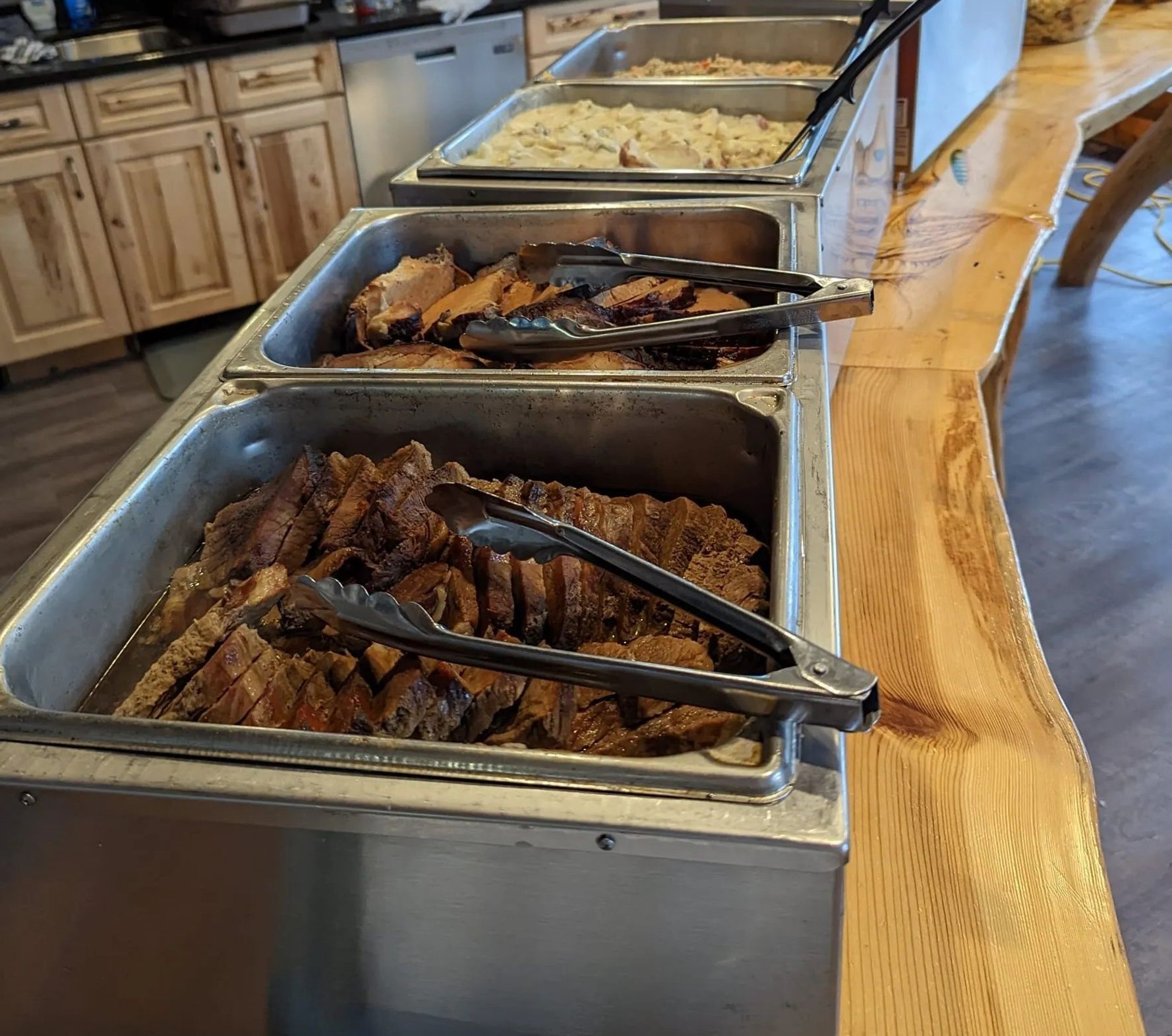 A buffet line with lots of food and tongs on a wooden table.